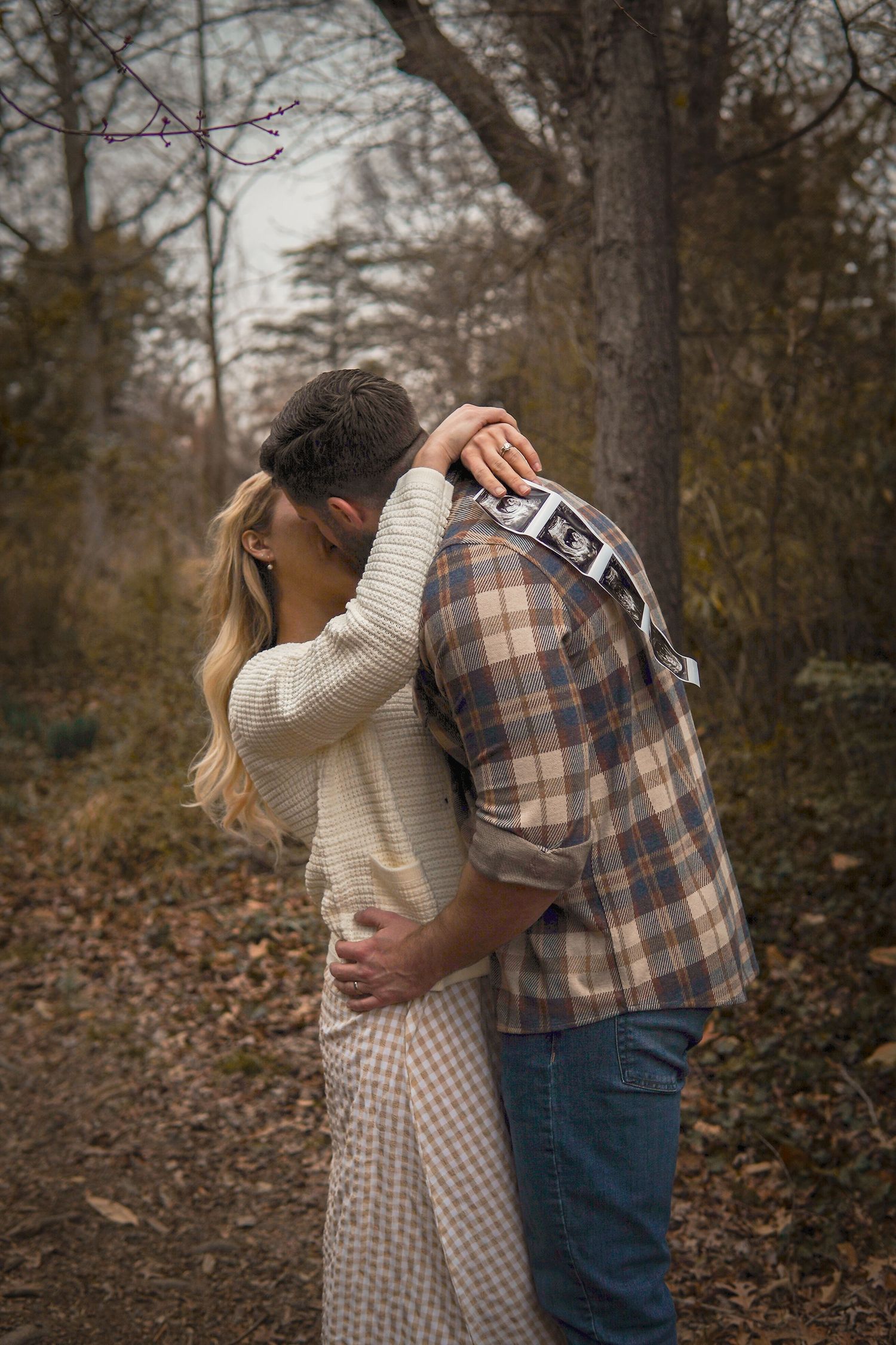 A couple embraces on a wooded autumn path, wearing a knit sweater and plaid shirt in muted earth tones.