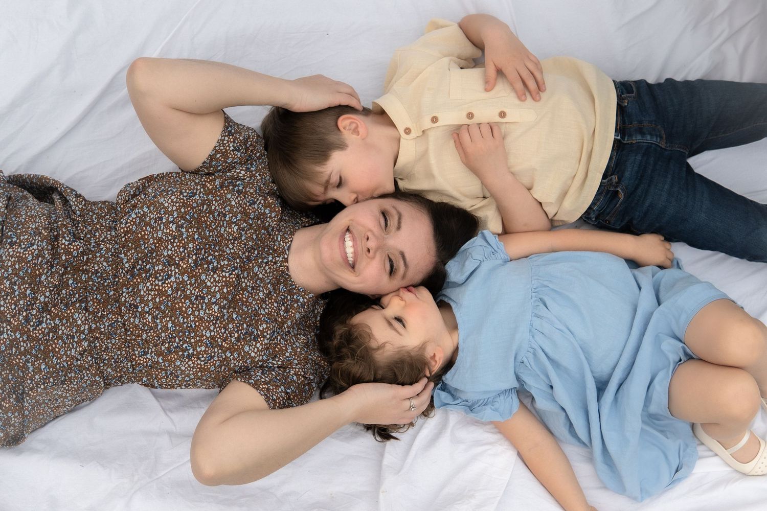 Three people lying close together on white bedding in casual clothing looking relaxed and peaceful.