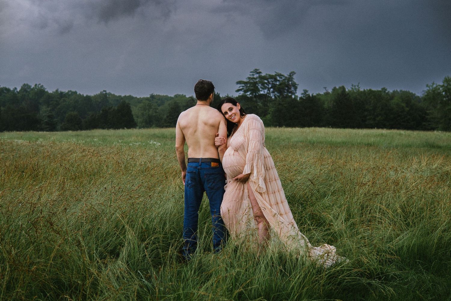 A couple stands together in a grassy field on a cloudy day with a tree line on the horizon.
