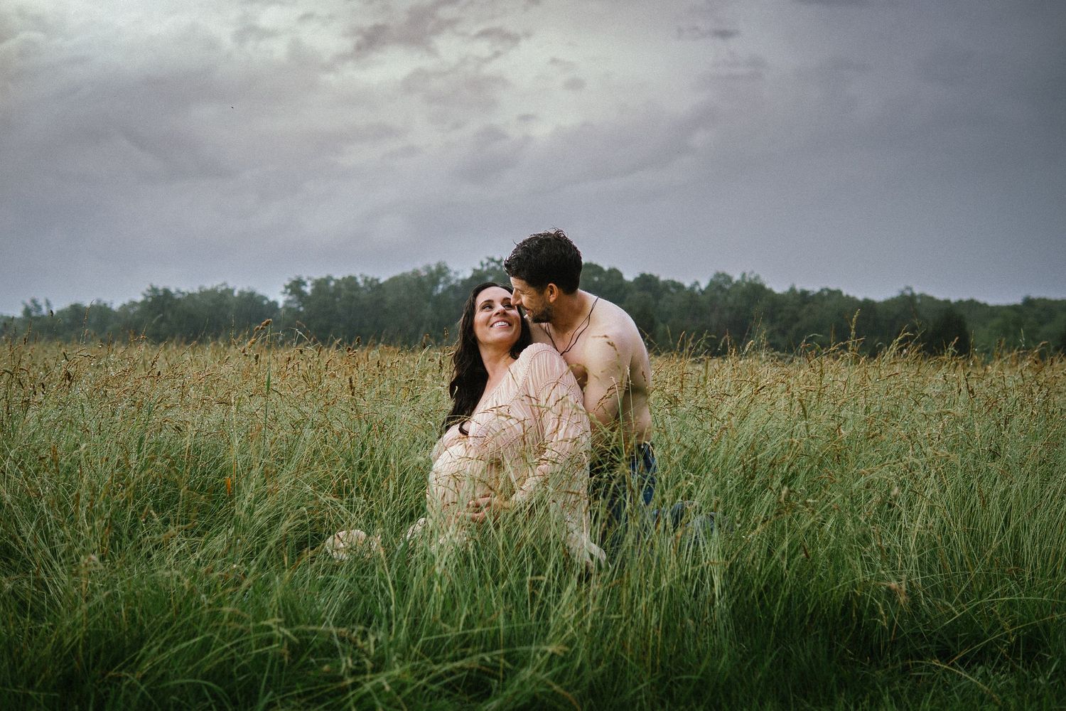 A couple embraces tenderly in a tall grass meadow under moody cloudy skies during sunset.