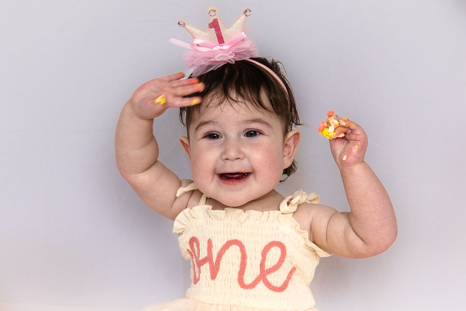 A baby wearing a pink crown and cream-colored outfit raises their arms while smiling against a light gray background.