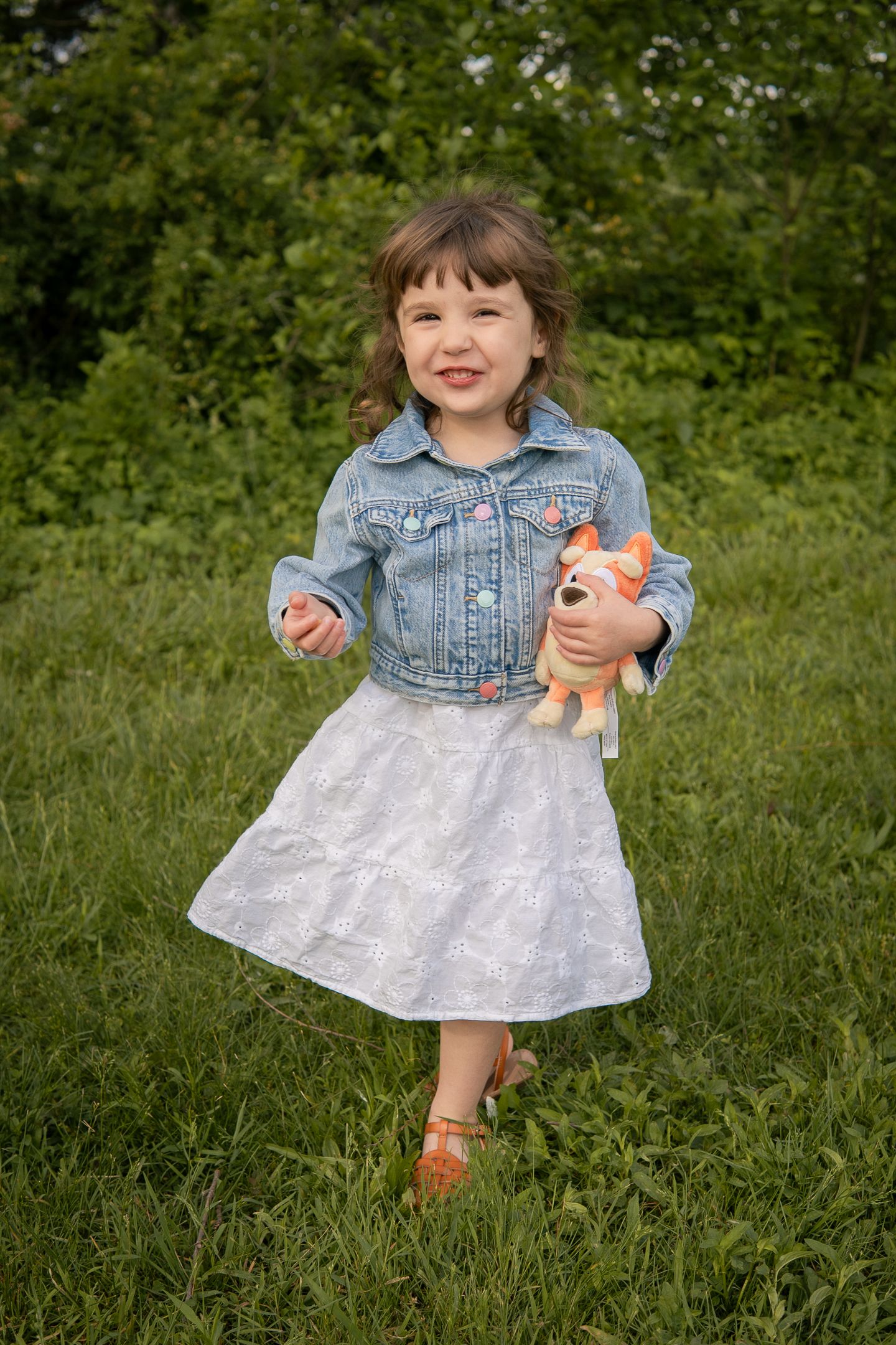 Child in denim jacket and white dress stands in green grass holding a stuffed animal toy while smiling.