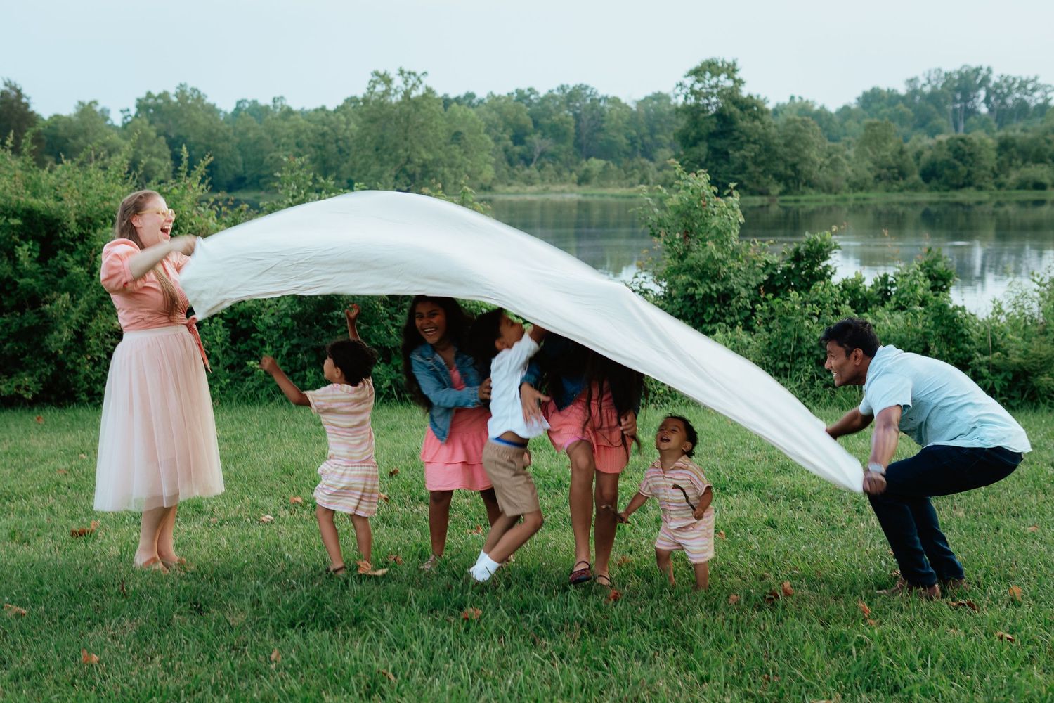 A playful wedding party runs through a grassy field near a lake while holding up the bride's veil on a sunny day.