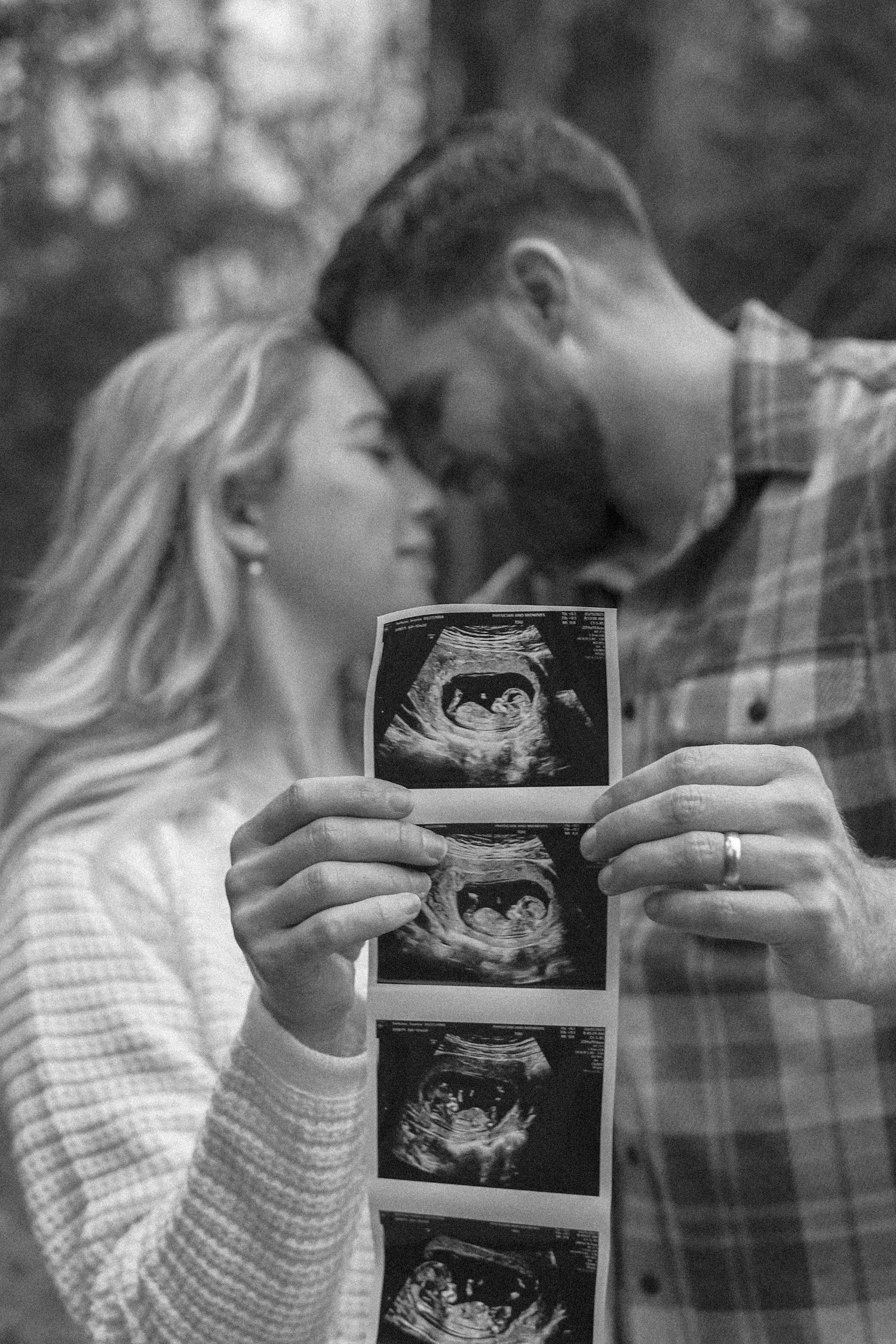 A couple holds up ultrasound photos while sharing an intimate moment in black and white photography.