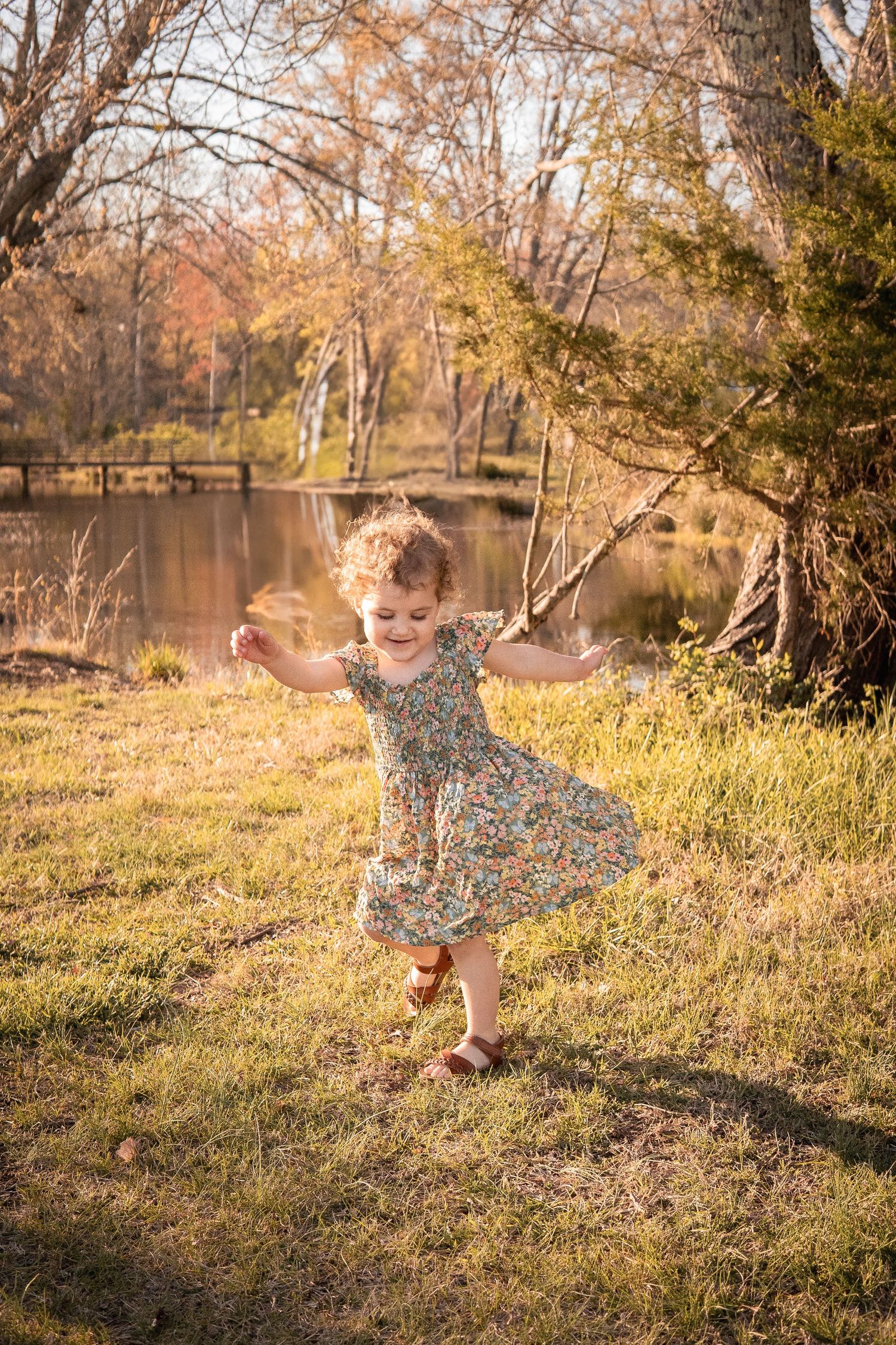 Child in floral dress dances joyfully near a scenic lake during golden hour as autumn trees frame the background in Haymarket, Virginia.