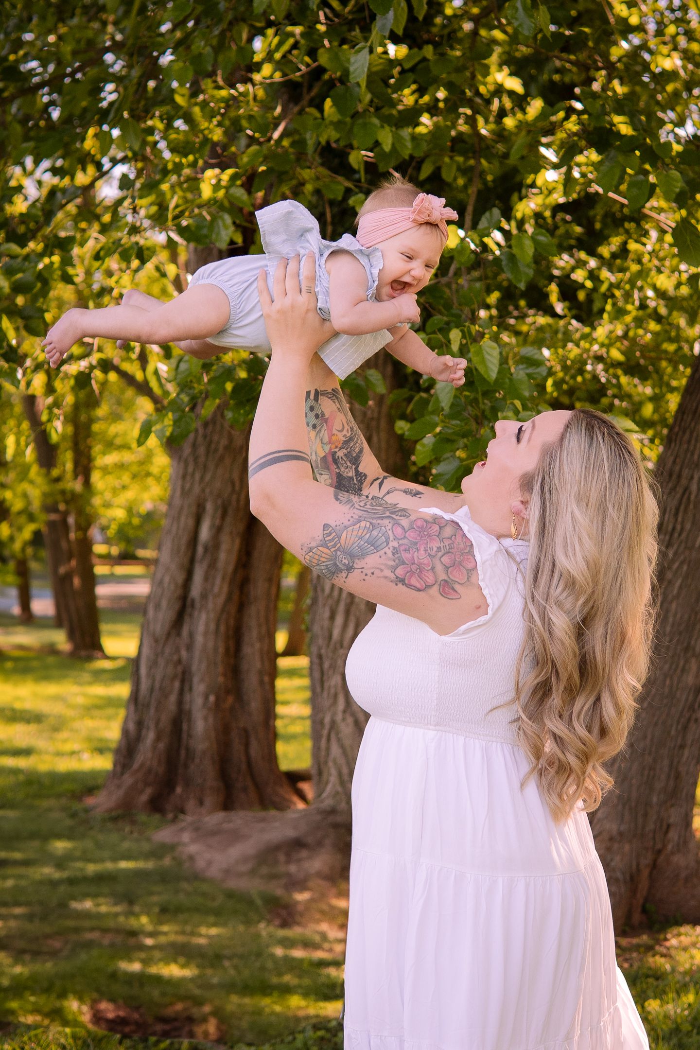 Mother lifts baby joyfully in outdoor park setting with row of trees in background.