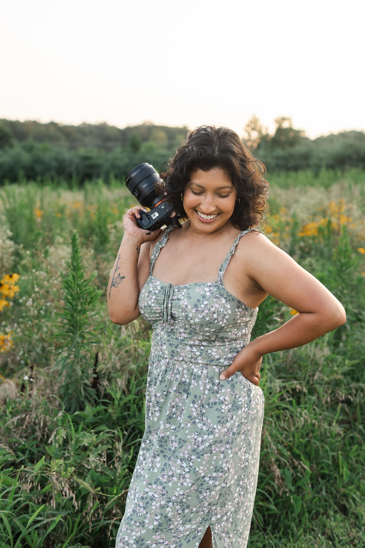 Woman in floral sundress poses joyfully with camera in wildflower field at sunset.