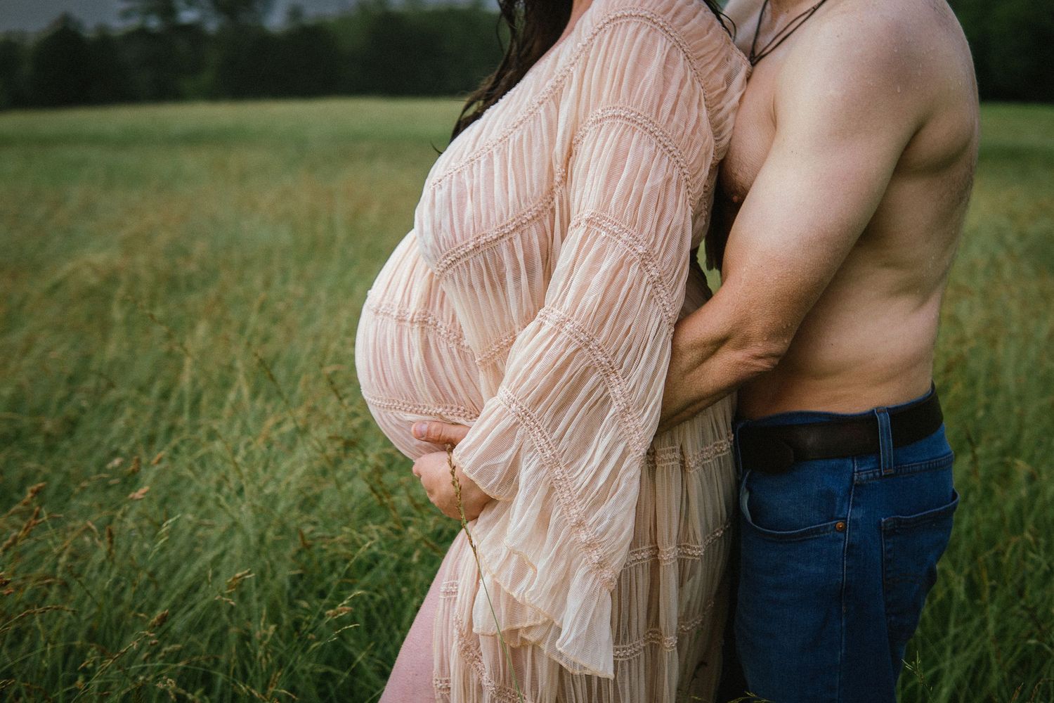 A couple embraces in an outdoor field wearing blue denim and flowing blush-colored fabric.