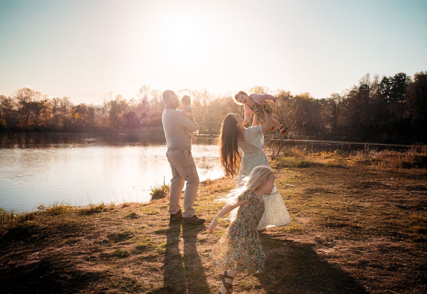 Family silhouettes holding hands and dancing together at sunset by a serene lake on a golden autumn afternoon in Haymarket, Virginia.