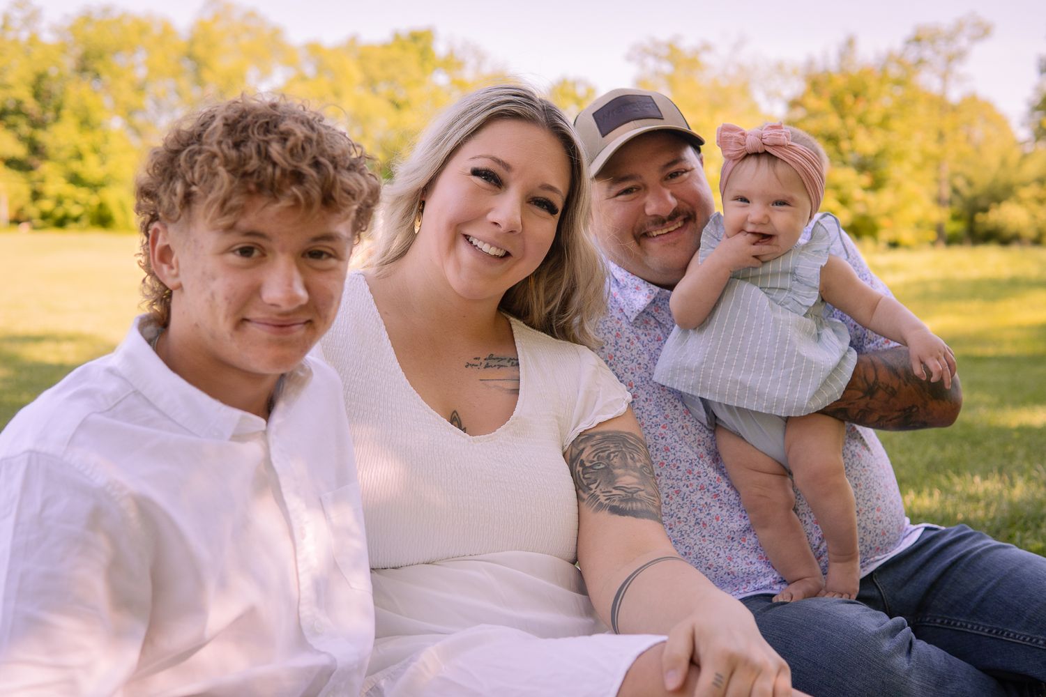 A family relaxes outdoors together on a warm afternoon in a grassy field with golden foliage behind them.