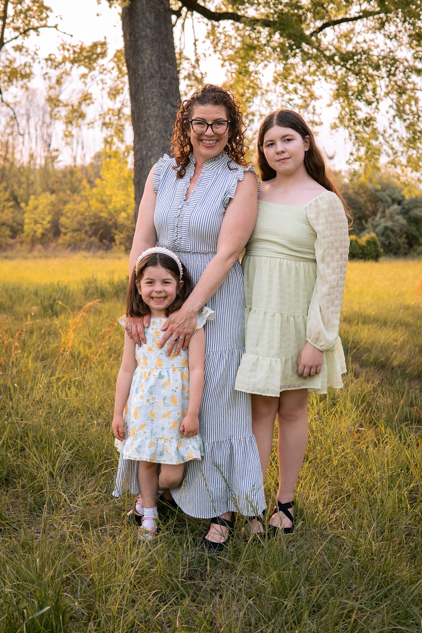 Three people pose together in summer dresses against a golden sunset backdrop in a grassy field.