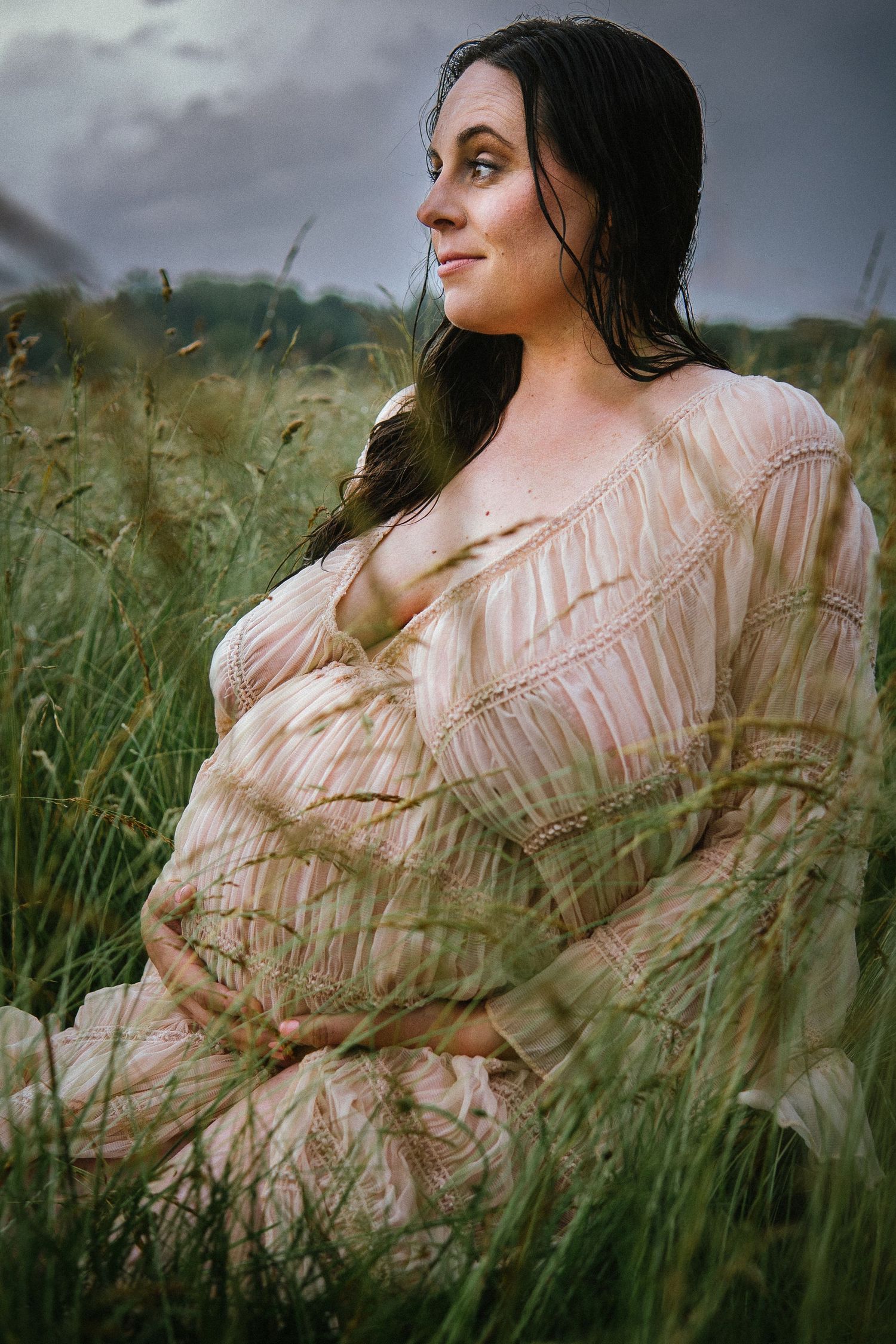 Woman in textured peach dress poses gracefully amid tall grass under moody sky.