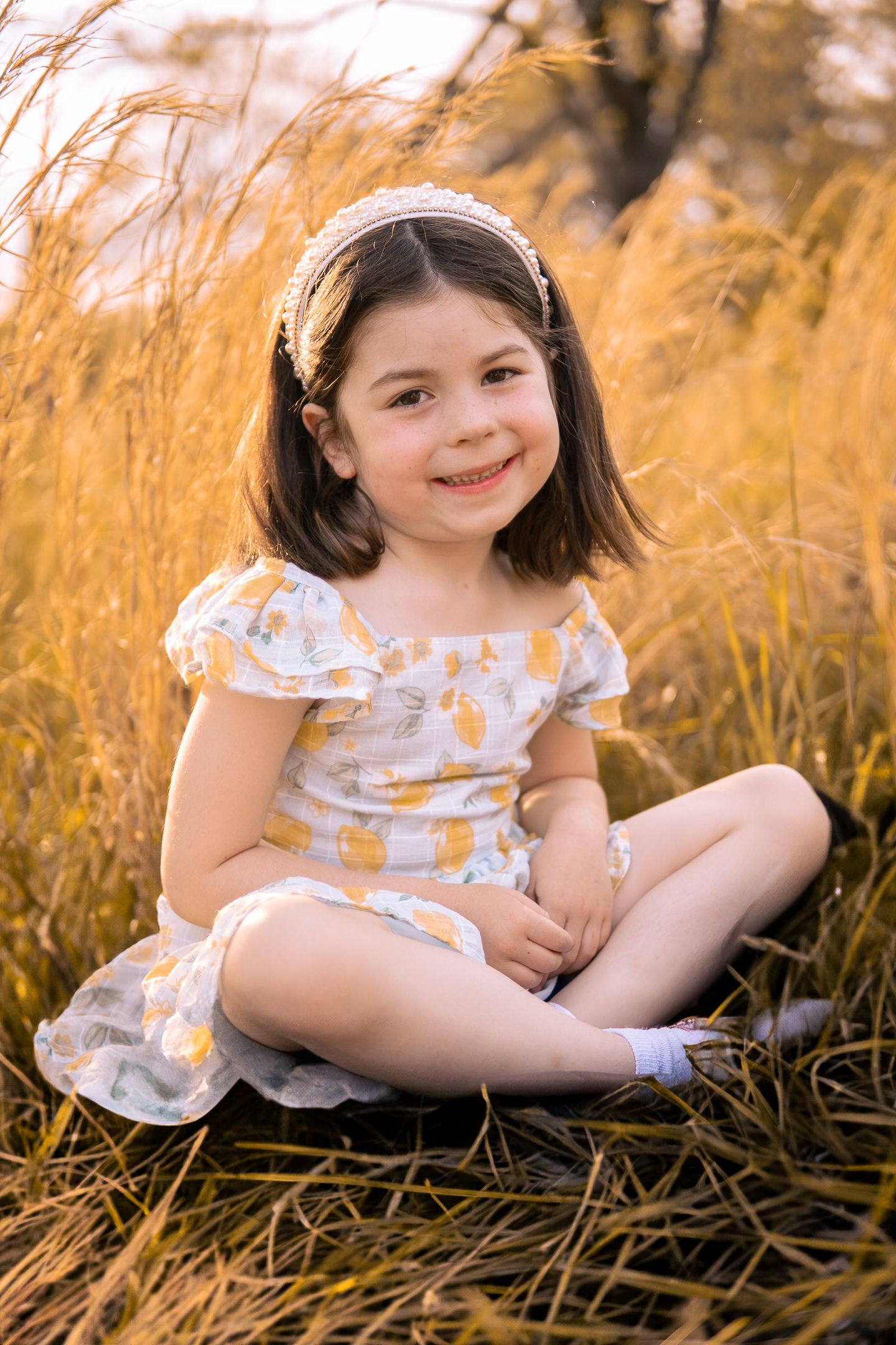 Young girl in floral dress sits in tall golden grass during sunset portrait session.