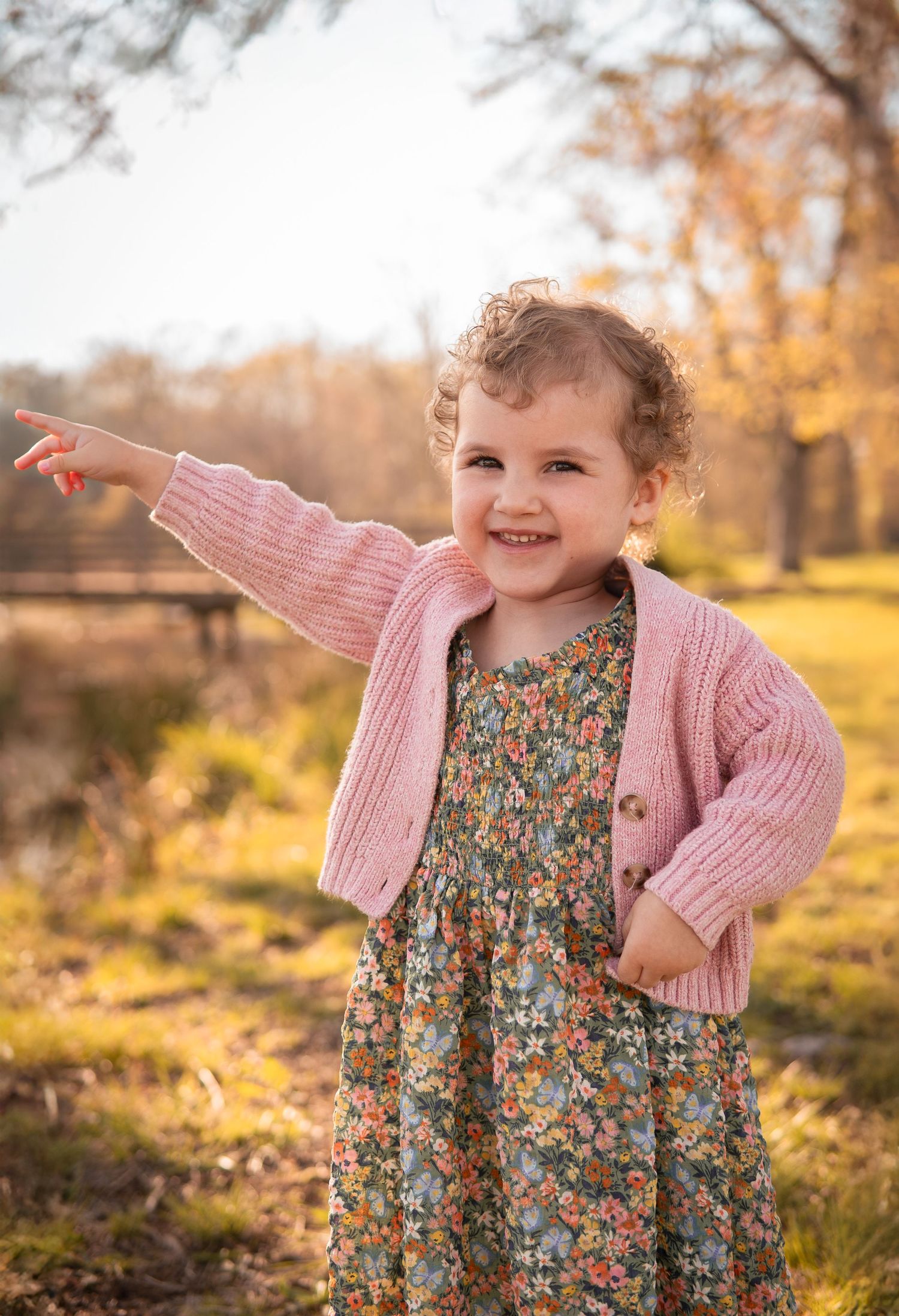 A joyful child in a pink cardigan and floral dress points while standing in a sunny autumn park setting.