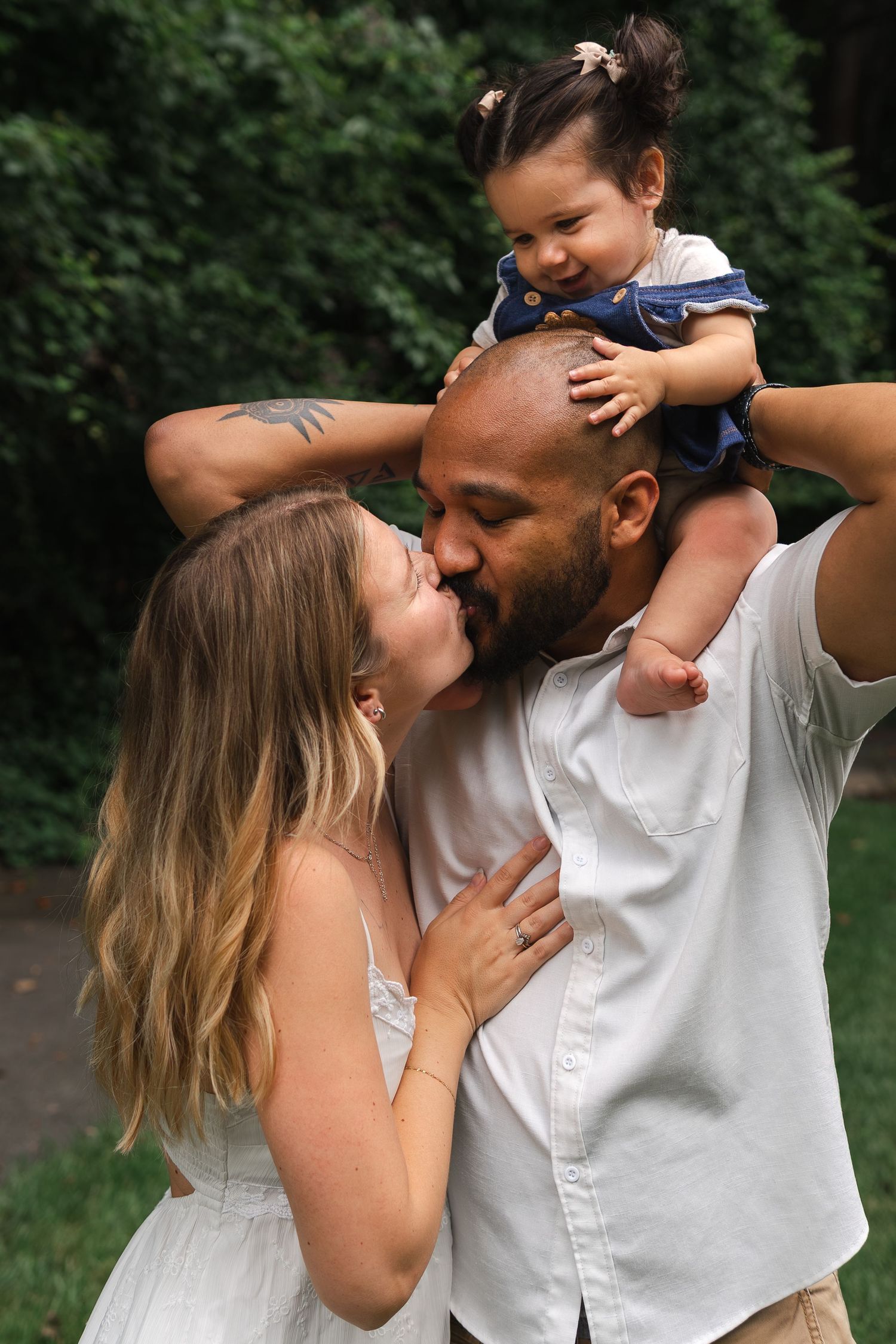 Loving family moment outdoors with parents embracing while holding a smiling baby wearing blue on shoulders.
