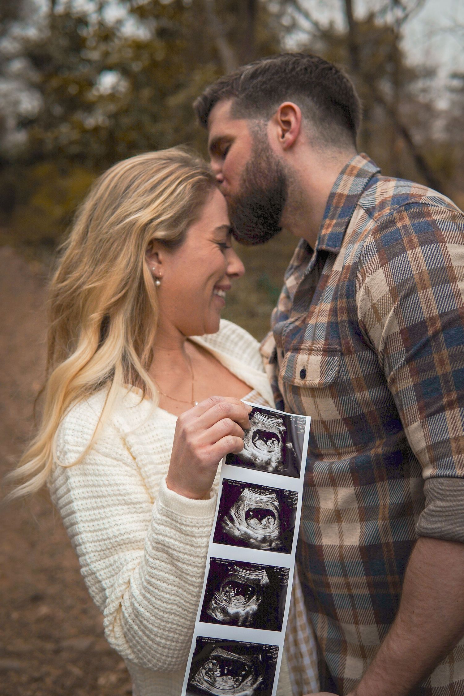 A couple shares an intimate moment while holding ultrasound photos in an outdoor fall setting.