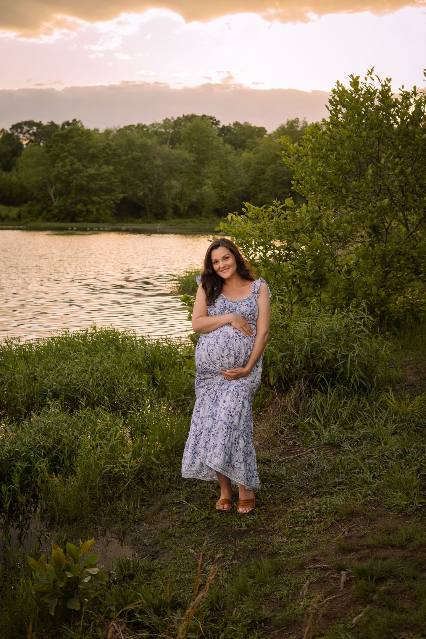 Sunset maternity photo by a lake with trees and golden sky in the background.