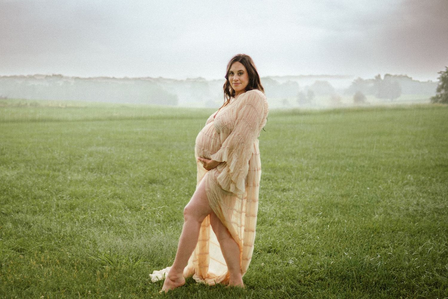 An ethereal maternity portrait on a misty green field shows an expectant mother in a flowing beige dress at sunrise.