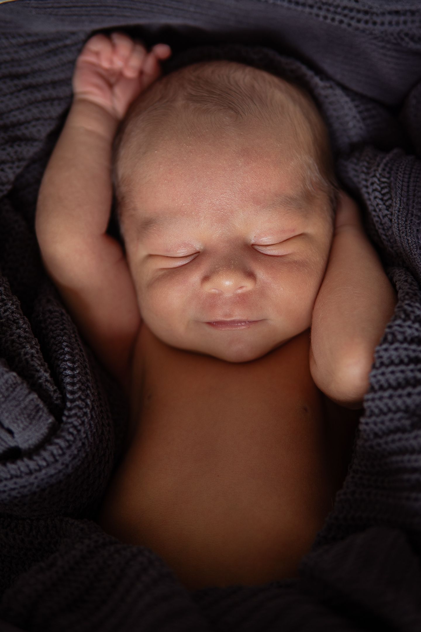 Peaceful newborn baby sleeping in soft dark knit blankets with one hand raised.