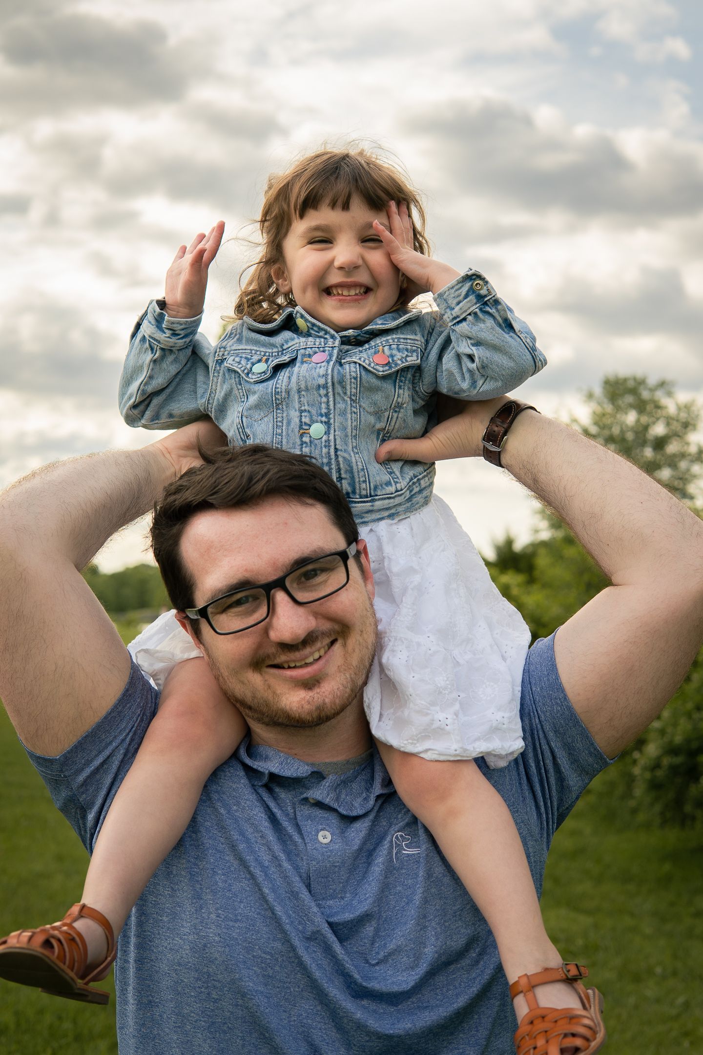 A joyful toddler in denim jacket sits on shoulders outdoors against a cloudy sky in a grassy field.
