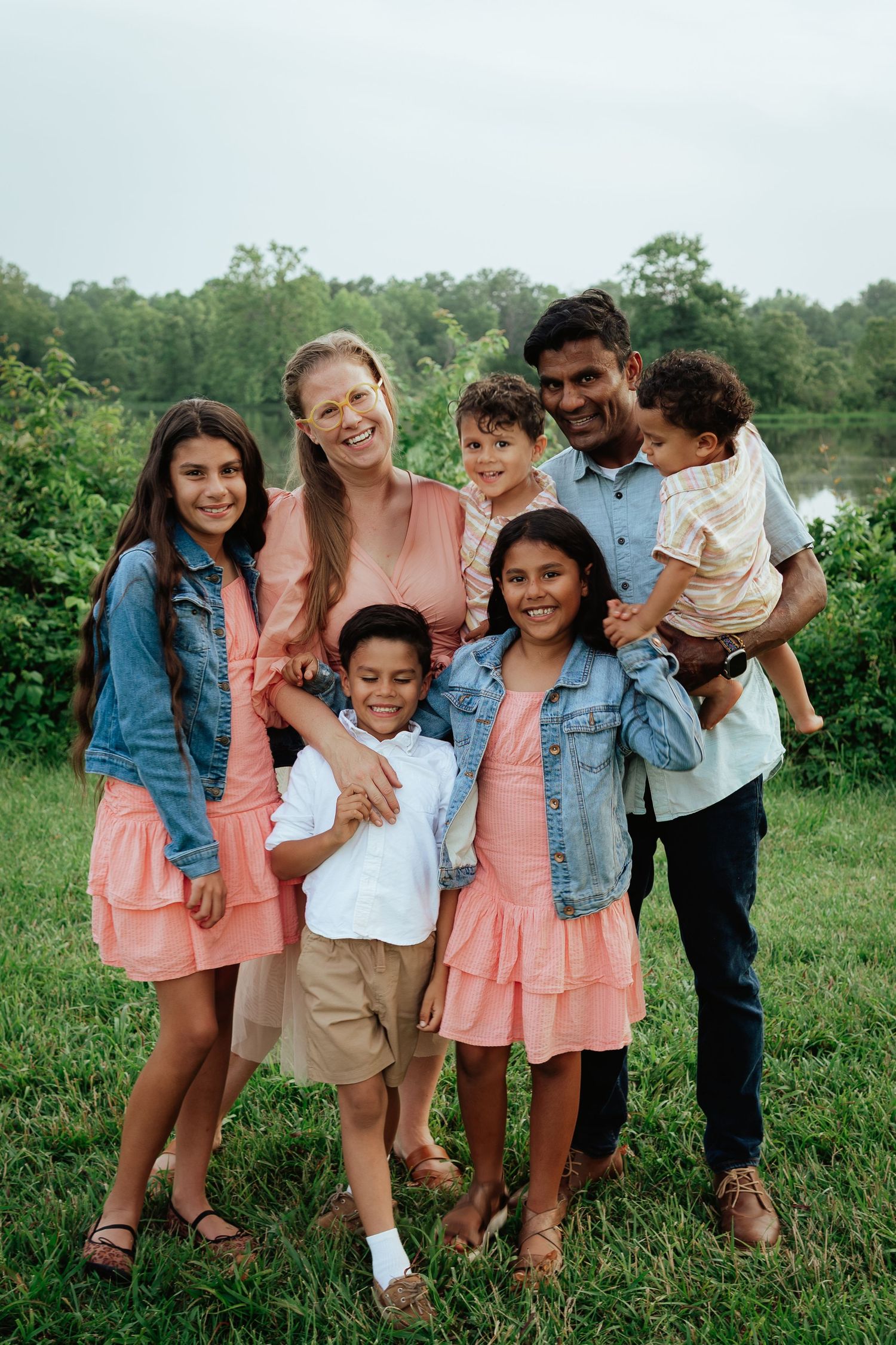 Happy family wearing coordinated pink and denim outfits poses together outdoors by a lake.