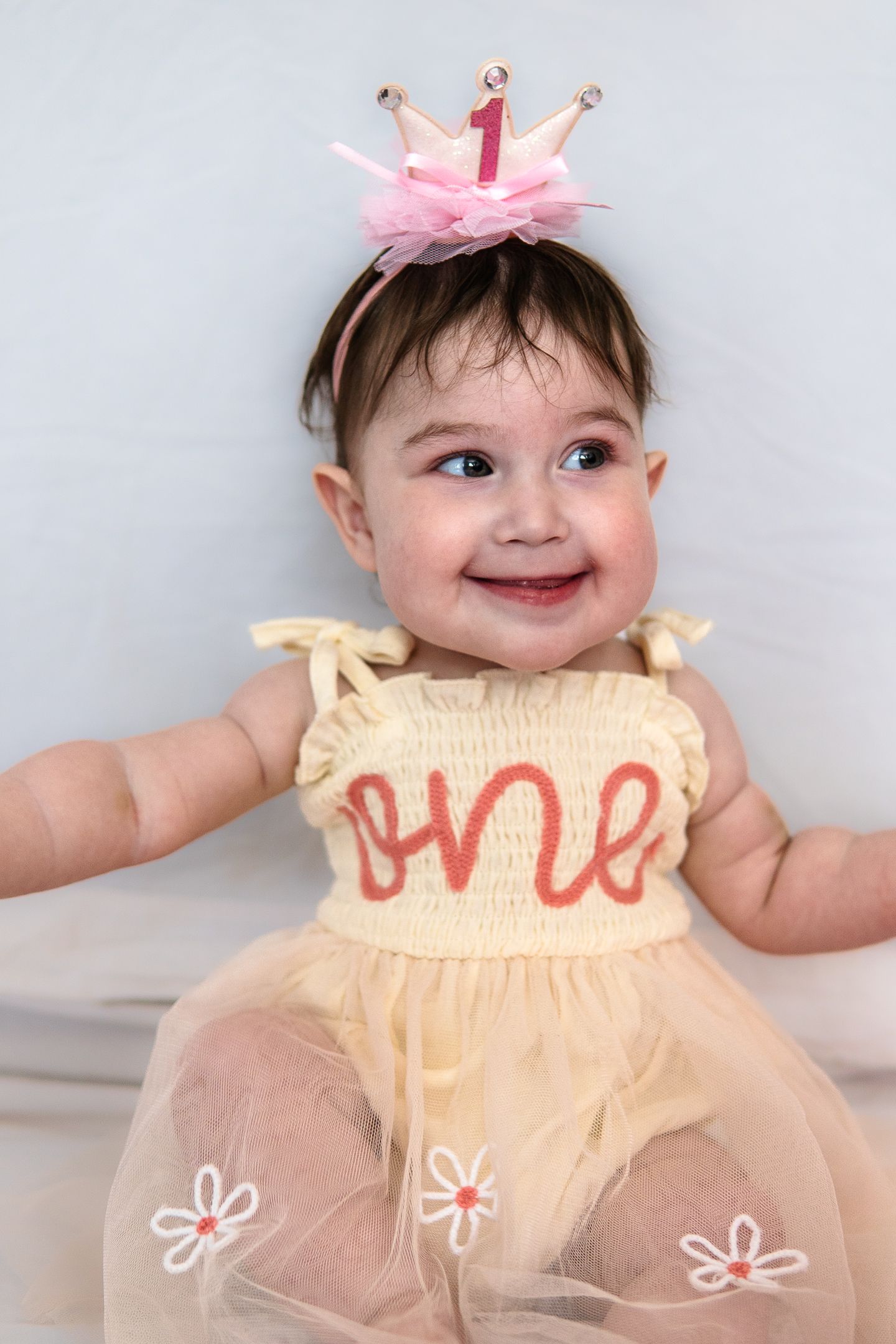 Baby wearing yellow tulle dress with pink crown accessory smiles at camera.