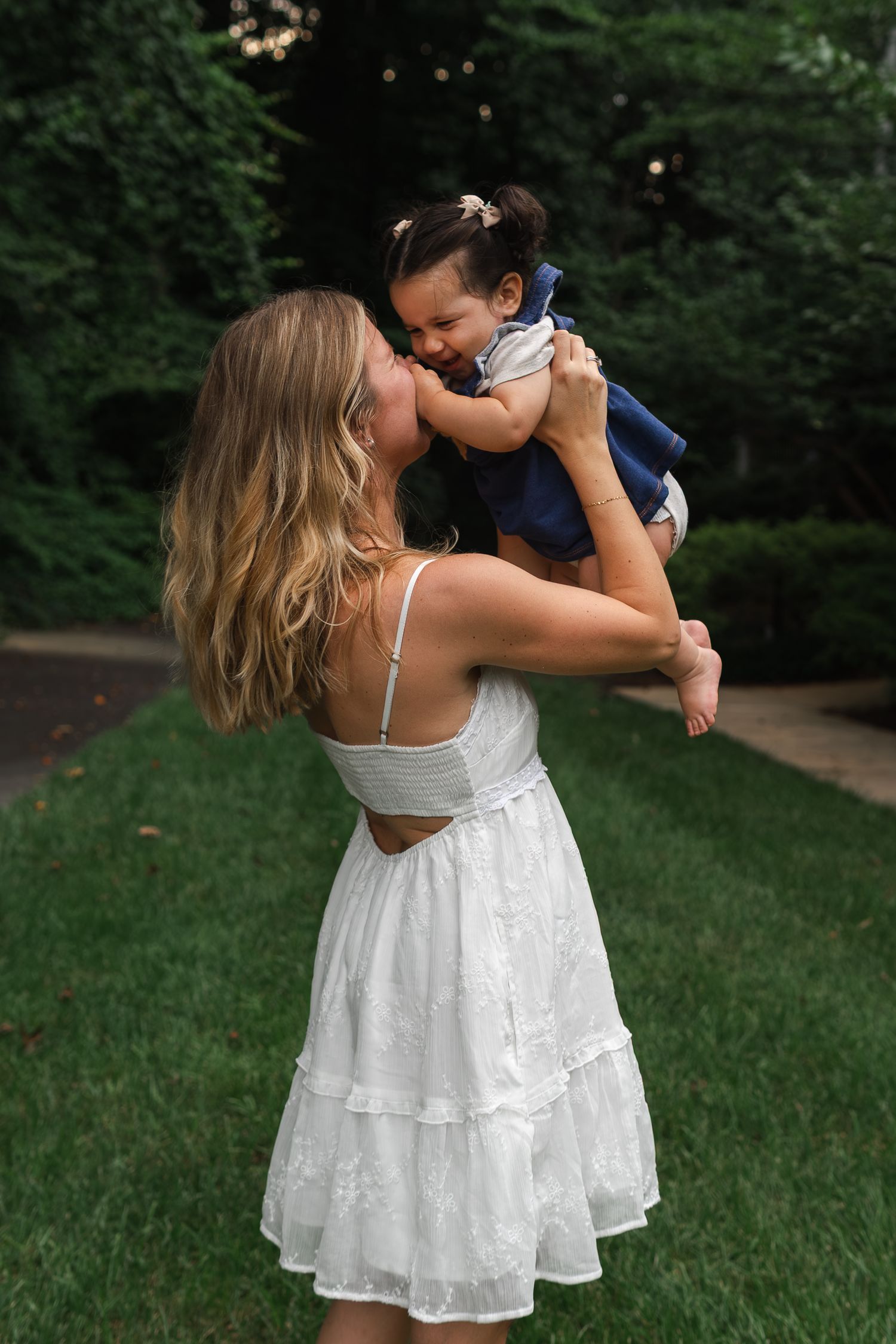A tender moment captured outdoors as a parent lifts a small child while wearing a white summer dress in a garden setting.