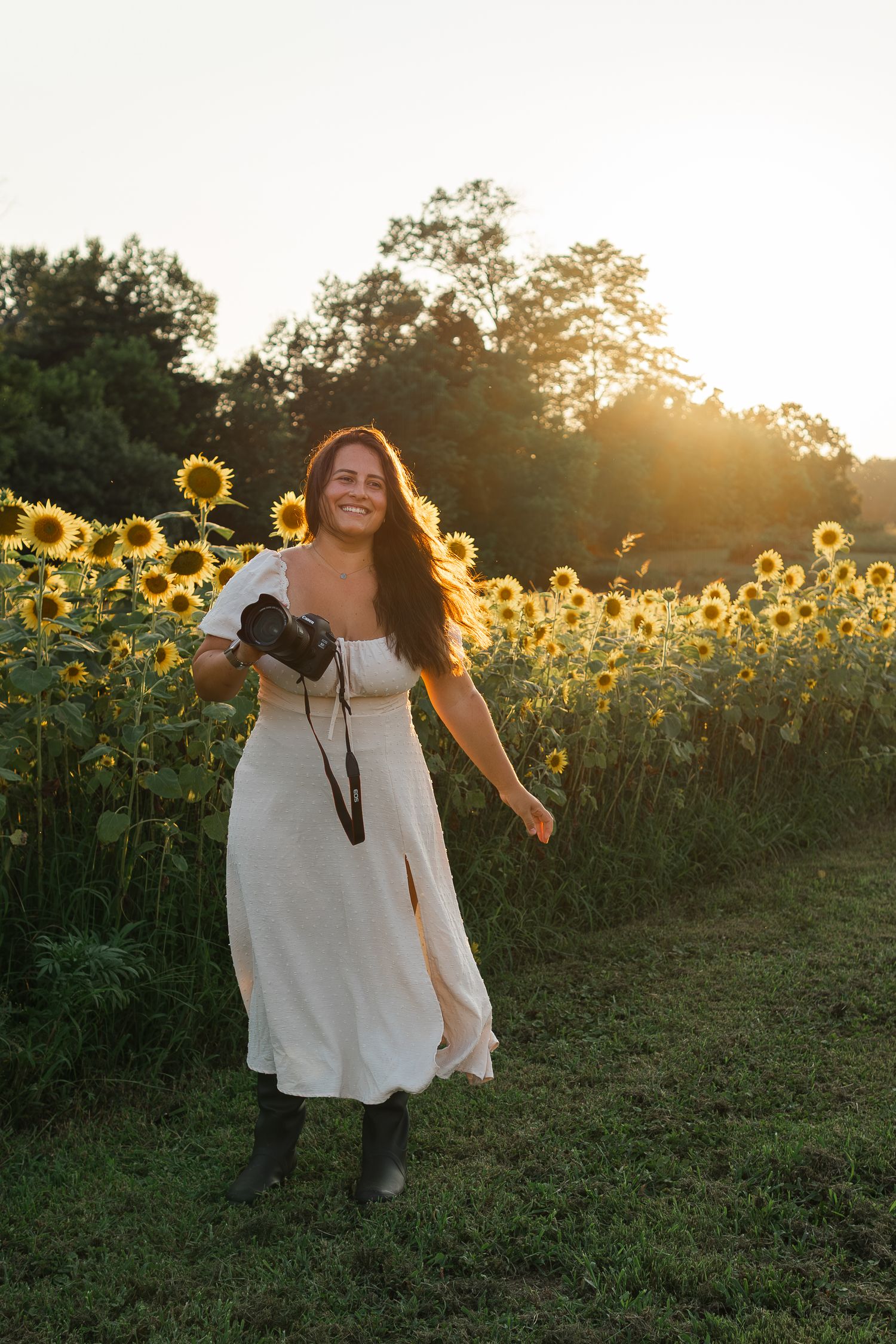 Photographer in white dress explores sunflower field during golden hour sunset with camera in hand.