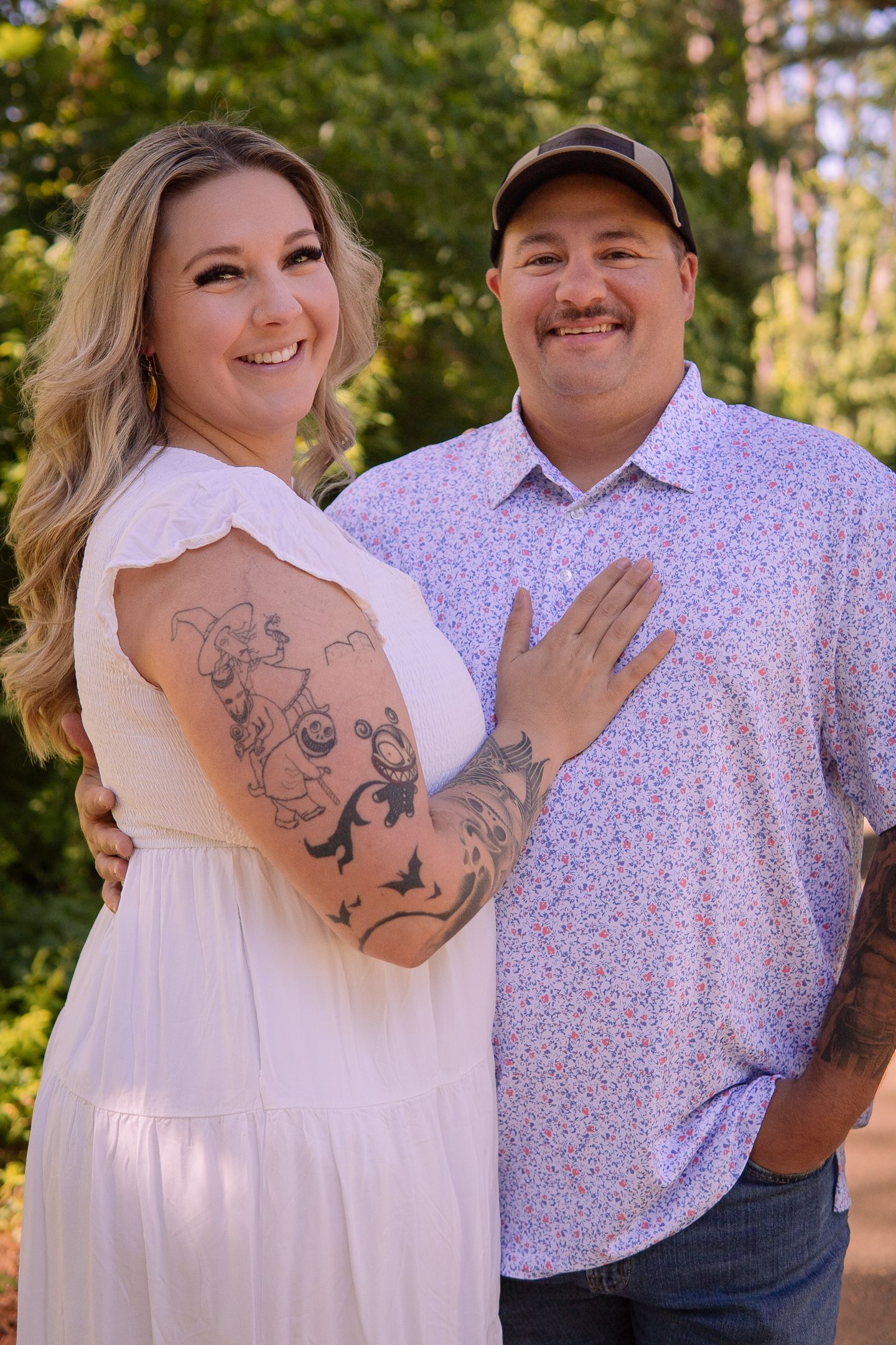 Couple in coordinating white and floral outfits pose outdoors showing artistic arm tattoos.