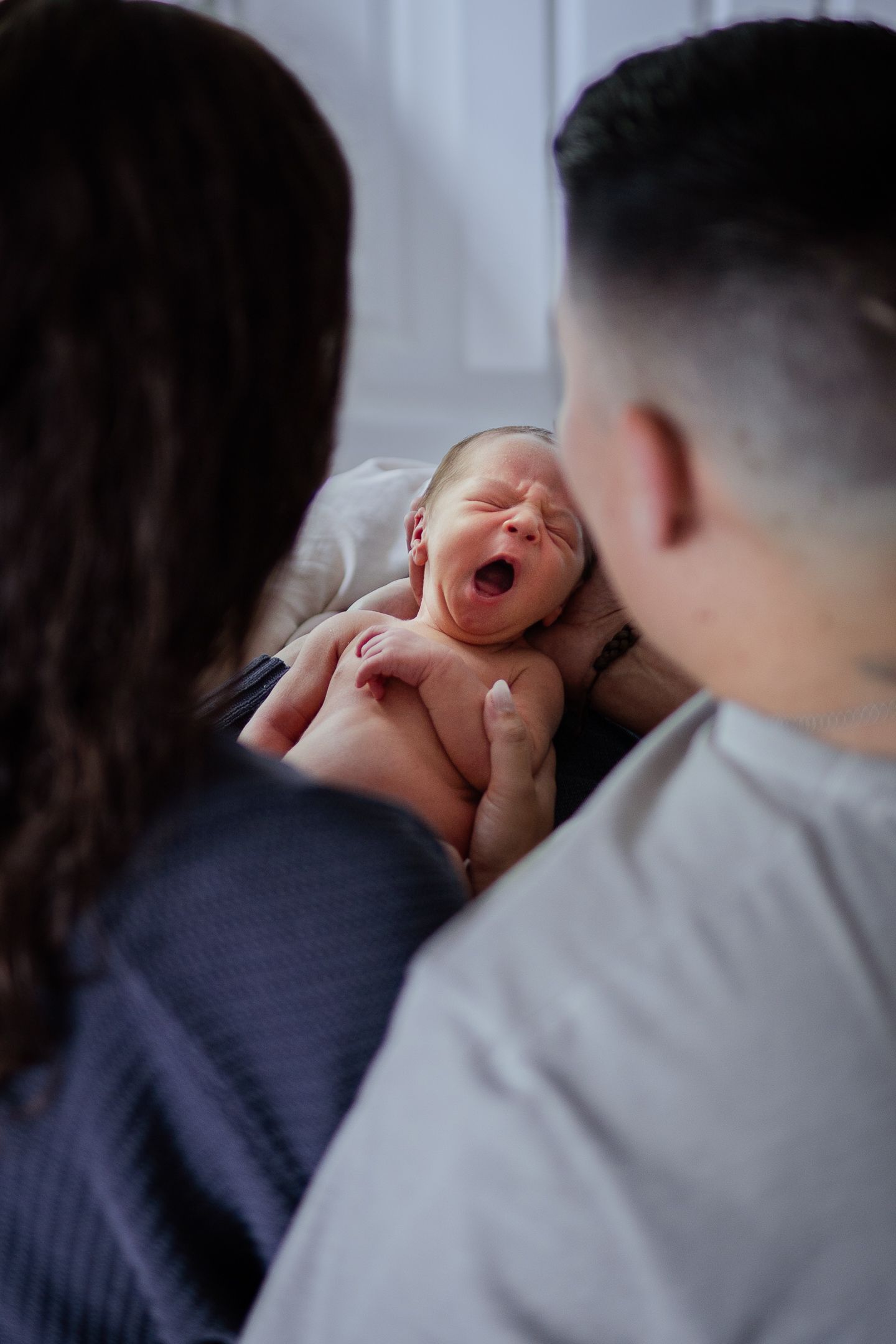 A newborn baby yawns while being cradled by parents in an intimate indoor portrait session.