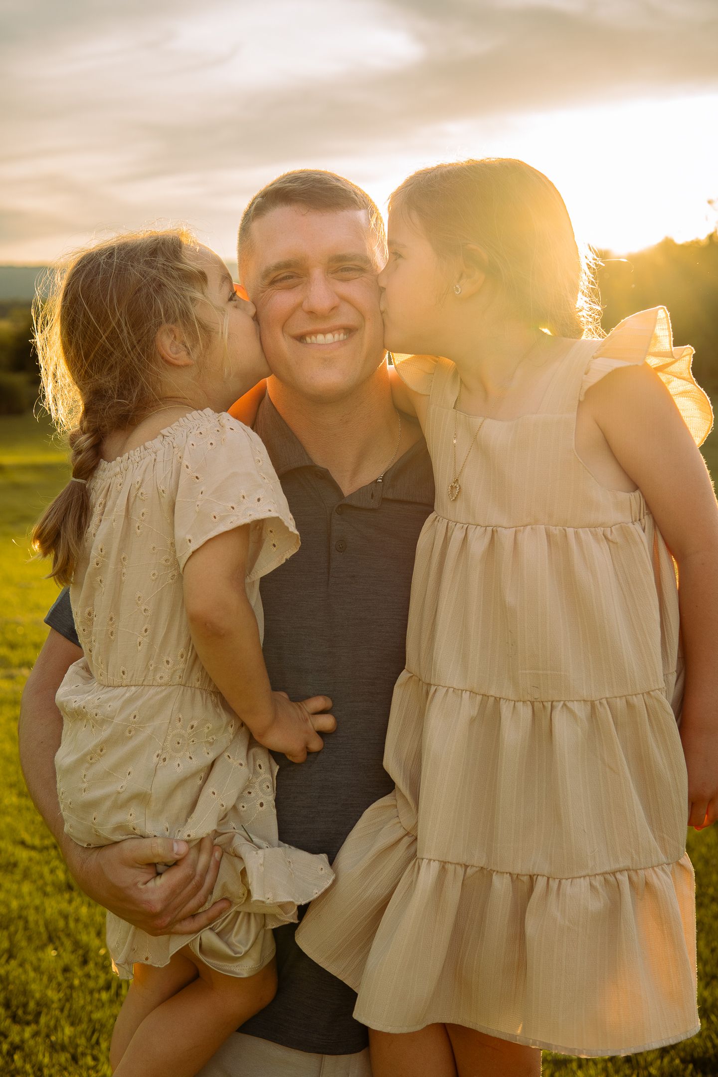 Two young children in light dresses give kisses on the cheeks during sunset in a grassy field.