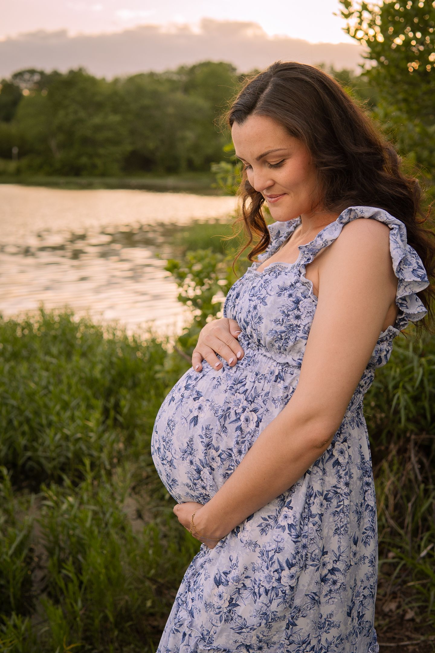 A pregnant woman in a blue floral dress stands by a lake at sunset, lovingly touching her belly.