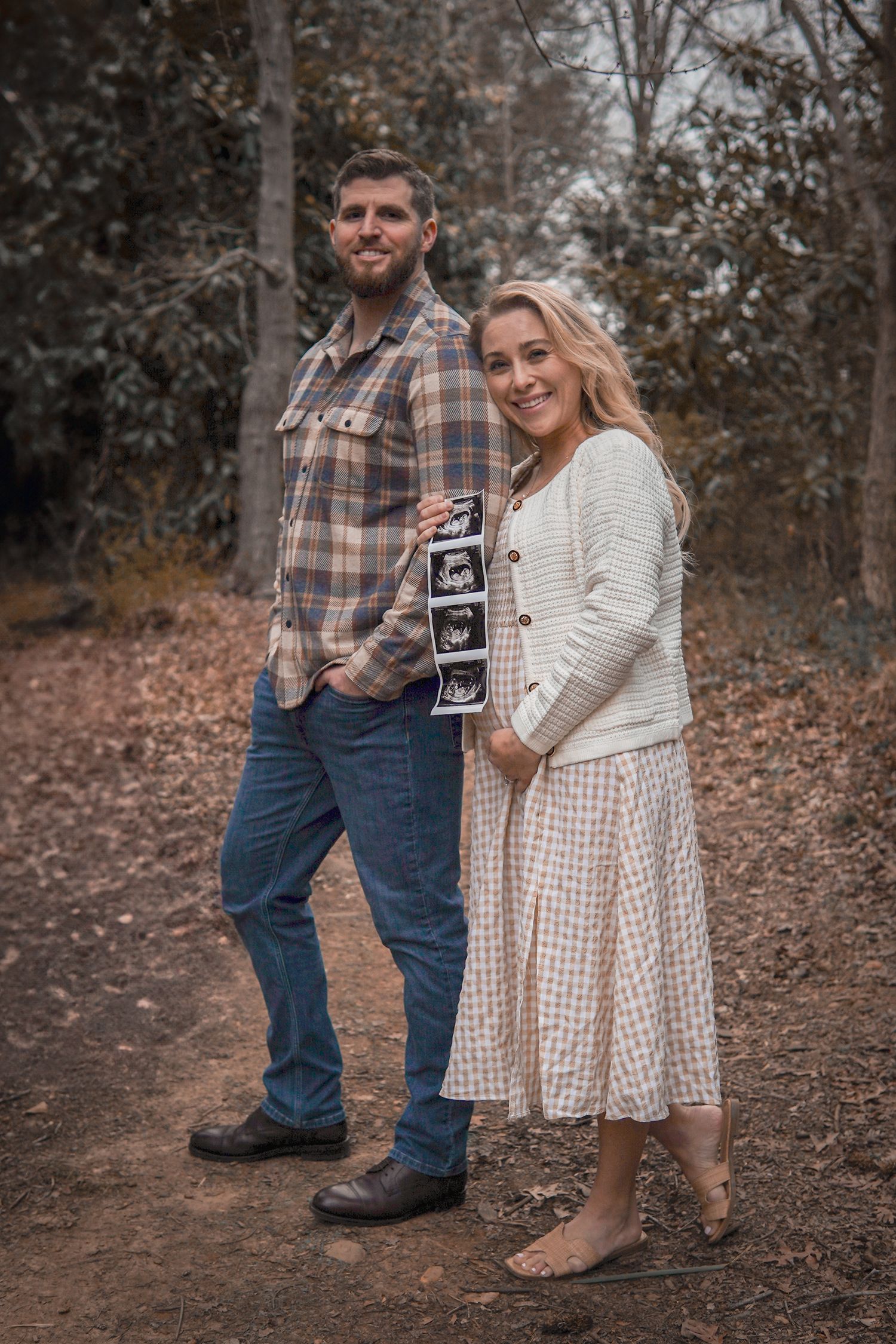 A couple stands on a dirt path in autumn woods holding ultrasound photos while announcing a pregnancy.