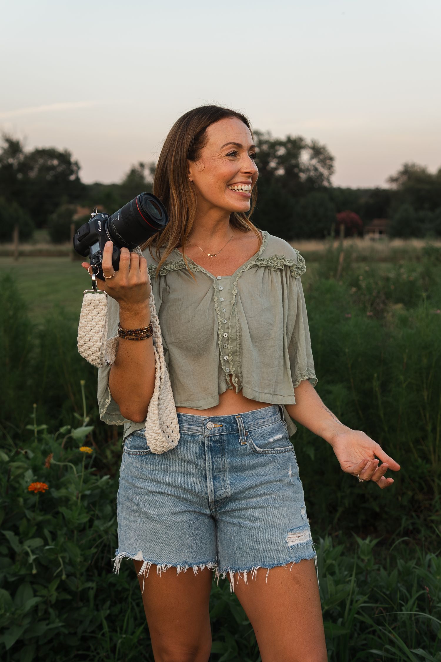 Photographer in casual summer outfit of crop top and denim shorts holds camera while laughing in field at sunset.