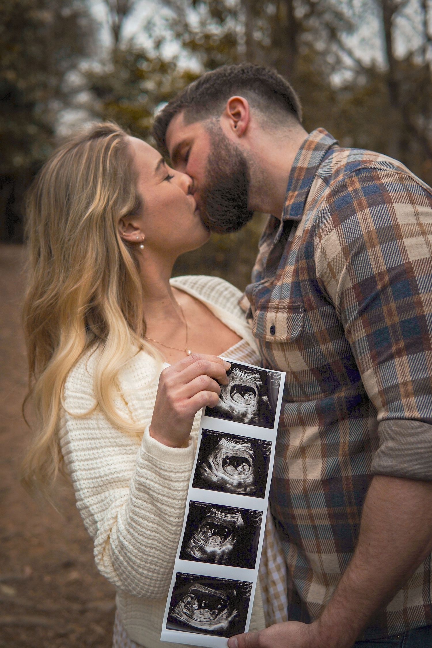 Couple shares intimate kiss while holding ultrasound photos in a romantic outdoor pregnancy announcement photoshoot.