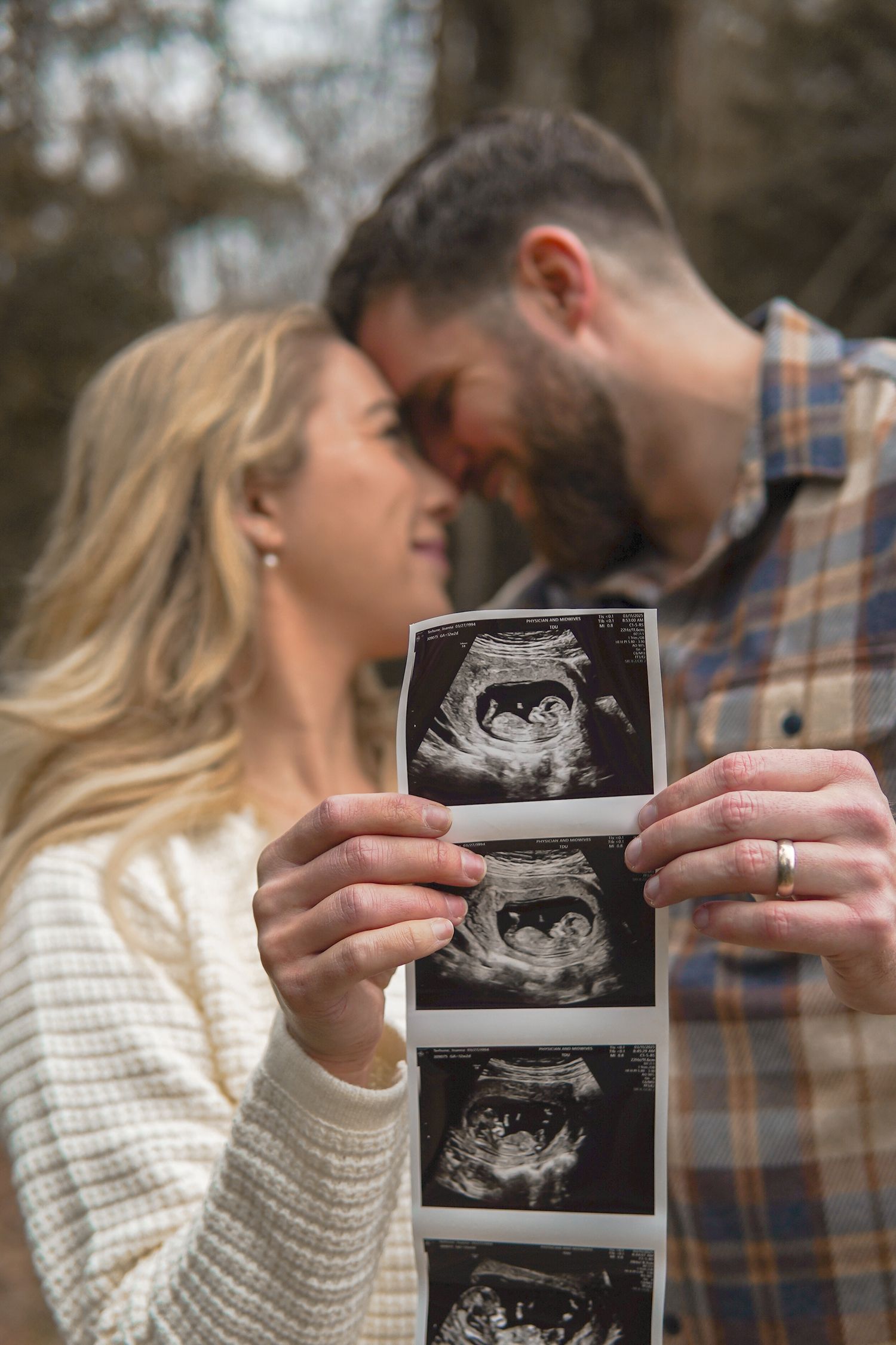 Happy couple sharing intimate moment while holding ultrasound photos of their unborn baby outdoors in nature.