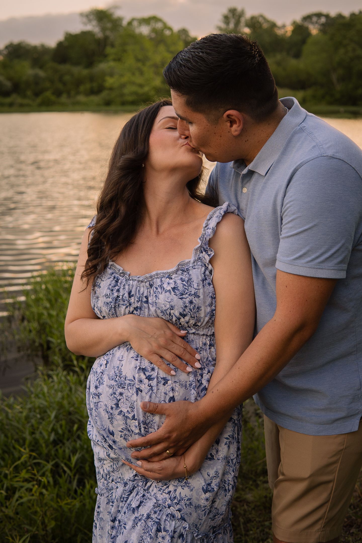 A romantic maternity photoshoot by a lake at sunset with a couple embracing in coordinated blue outfits.