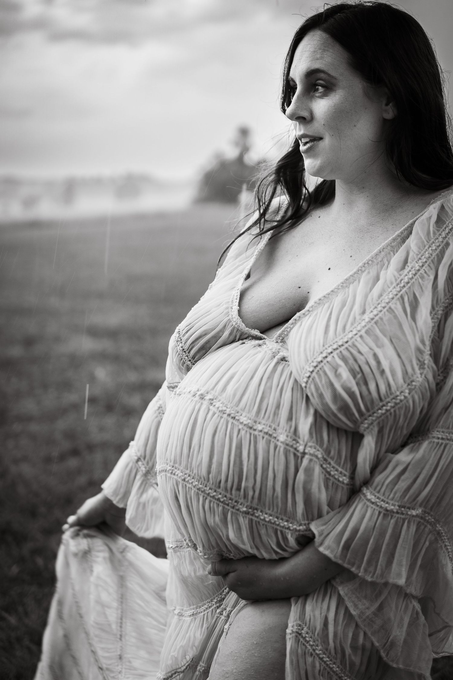 A dreamy black and white maternity portrait of a figure in a flowy white dress against a cloudy sky.