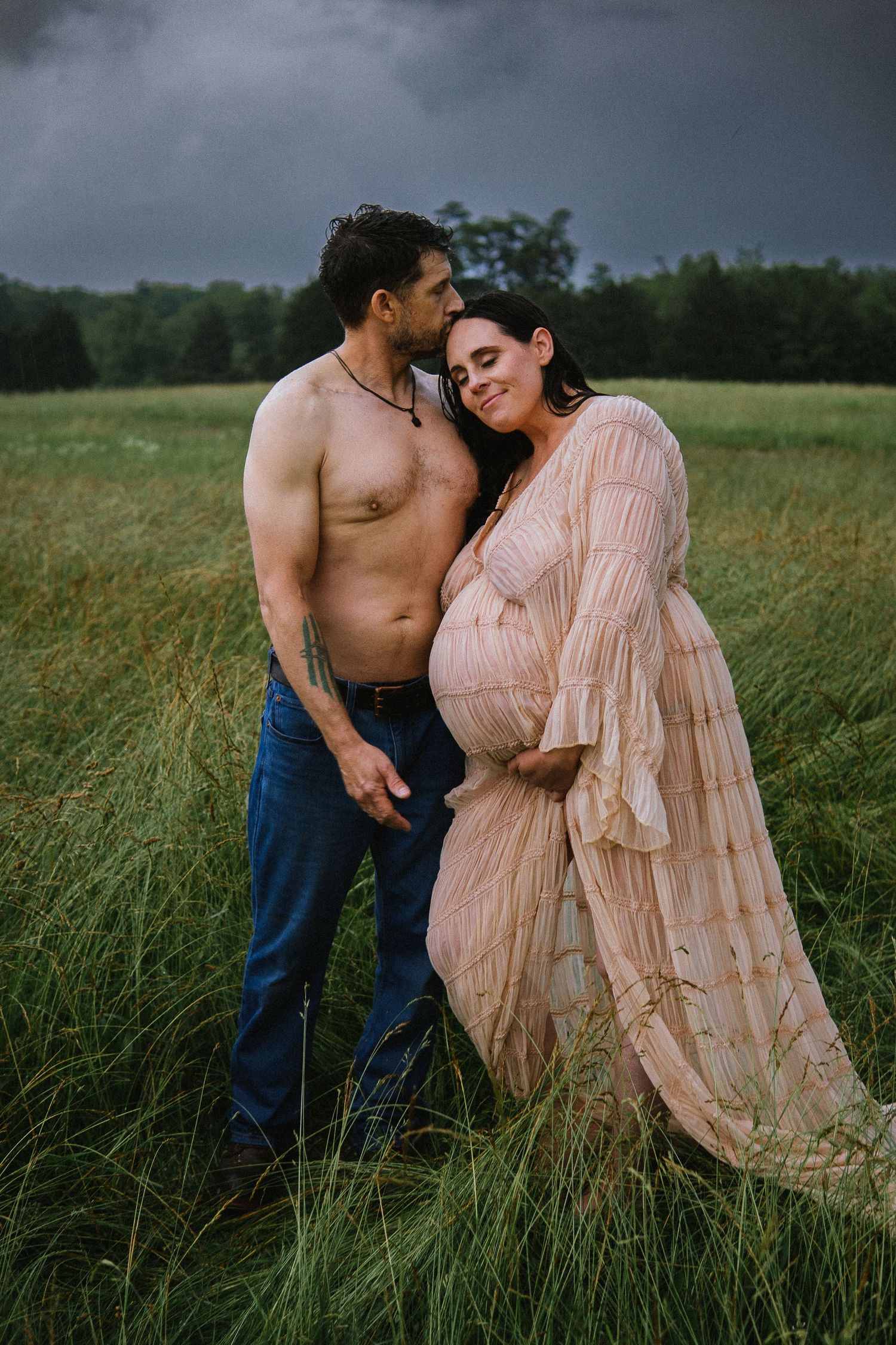 Romantic maternity photos of couple embracing in field with stormy sky backdrop.