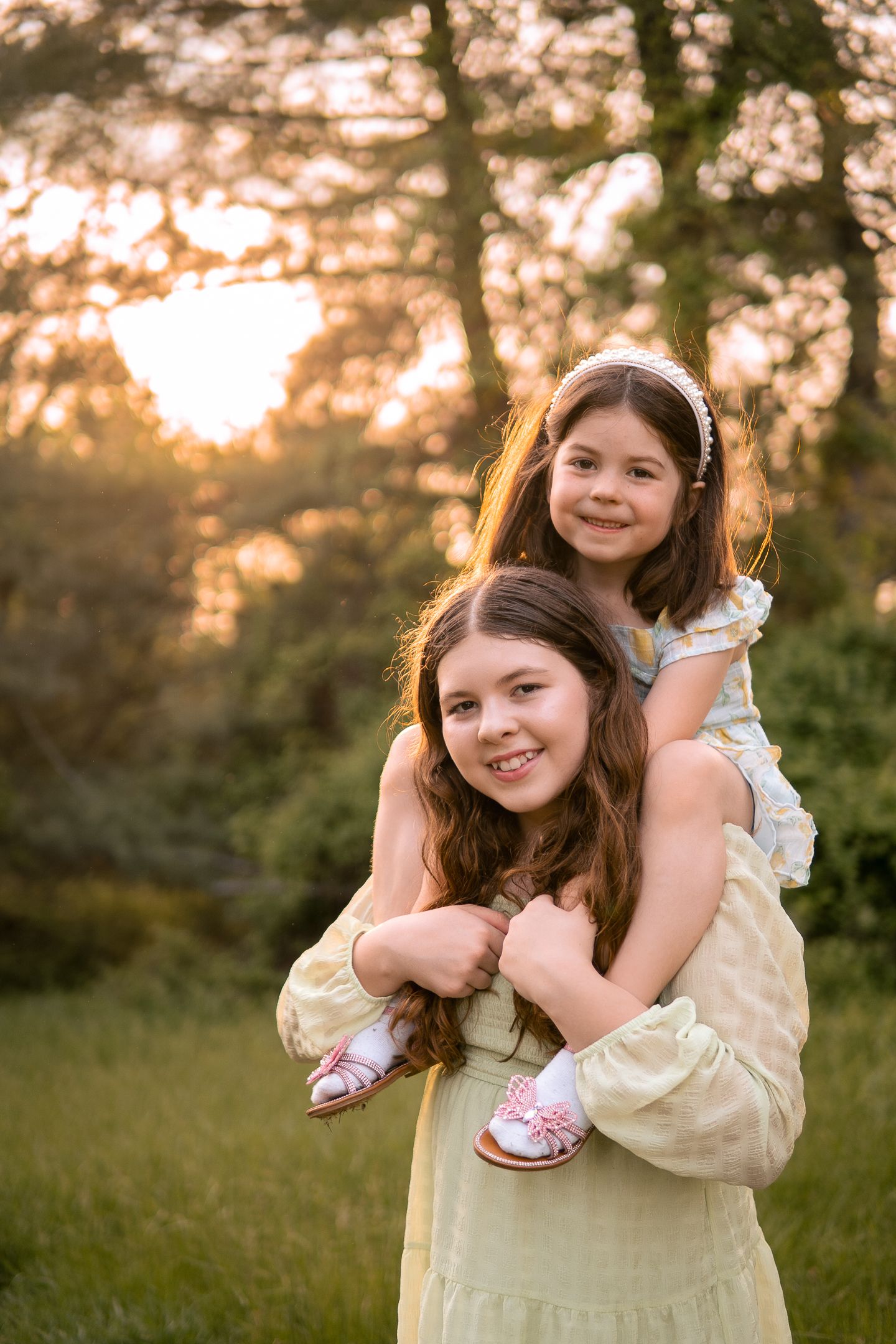 Two people embrace in a tender backlit outdoor portrait at sunset in a grassy area surrounded by trees.