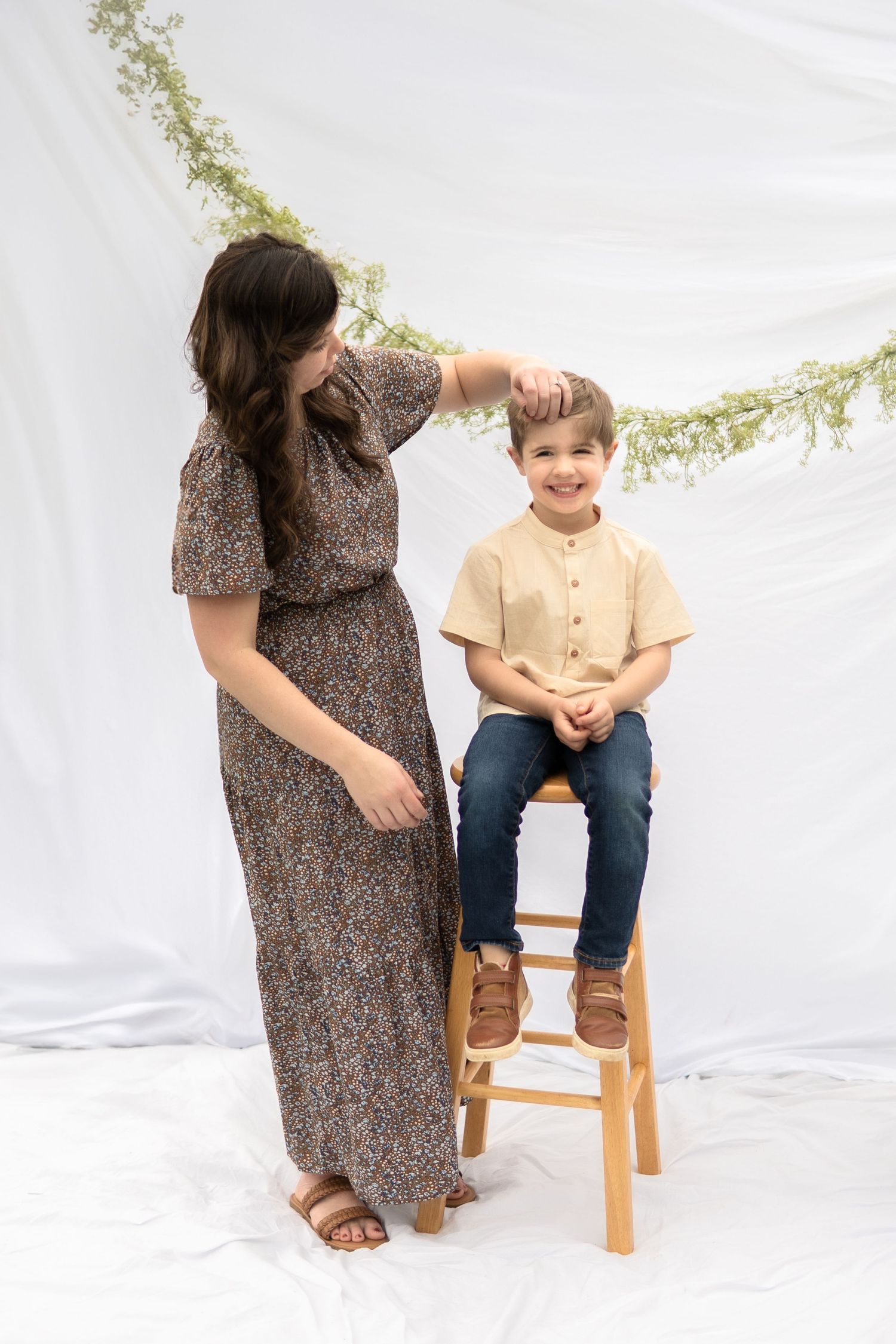 Person wearing floral maxi dress stands next to wooden stool against white backdrop with greenery garland.