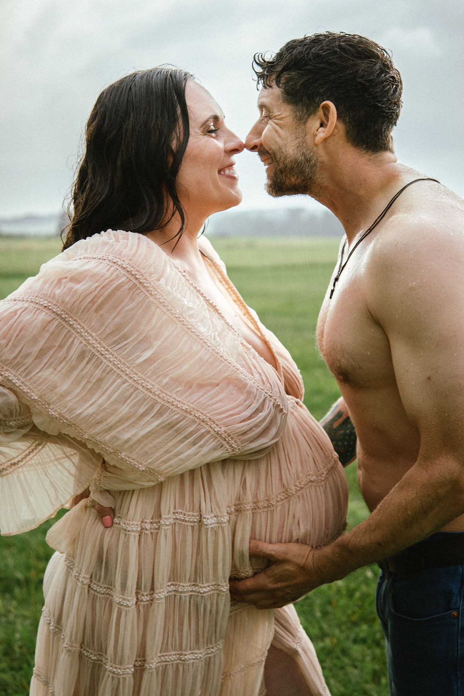 A romantic maternity photo shows a couple embracing in a field wearing soft pink and casual attire.