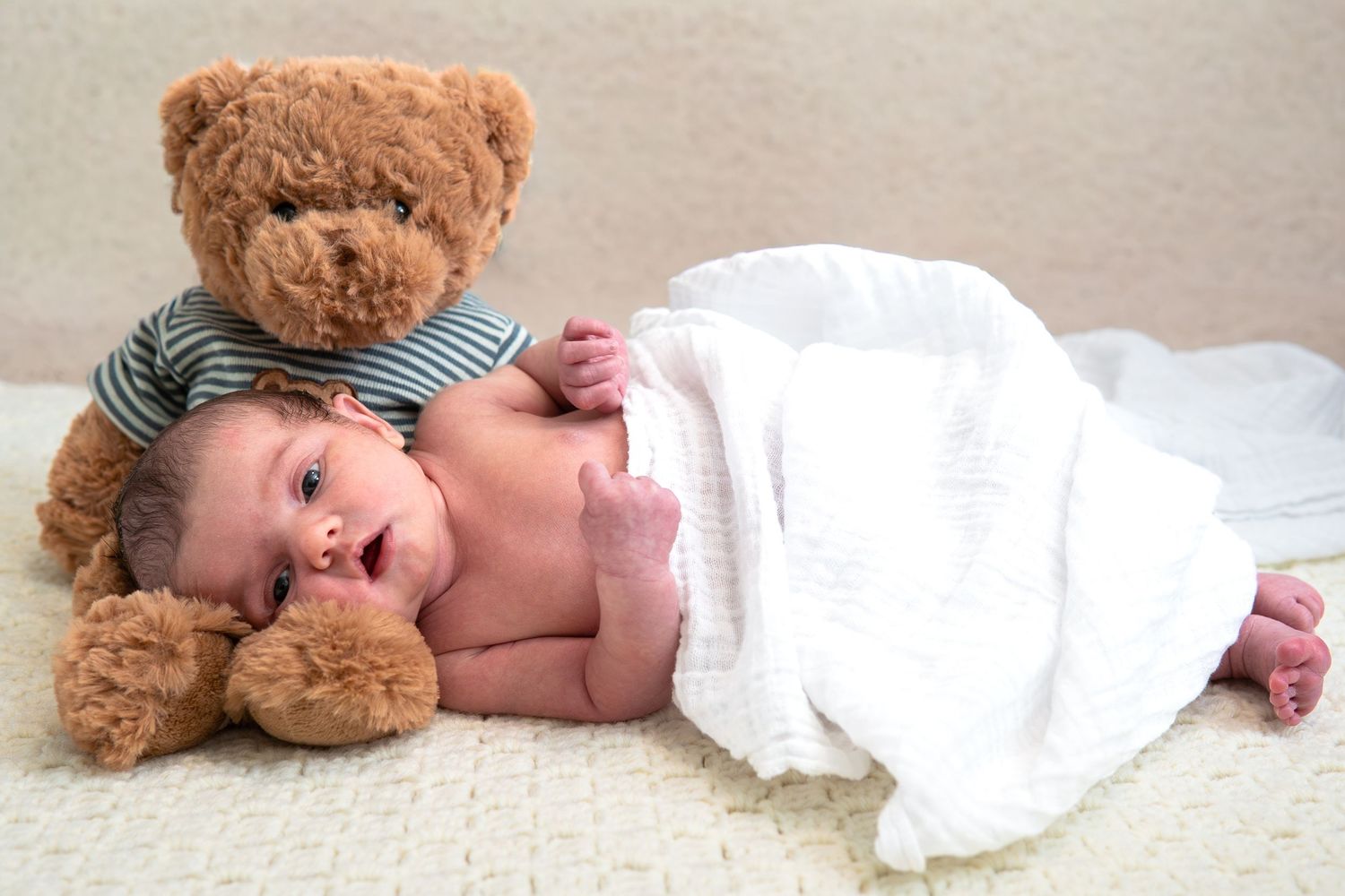 A sleeping baby lies on white fabric next to a brown teddy bear wearing a striped shirt.