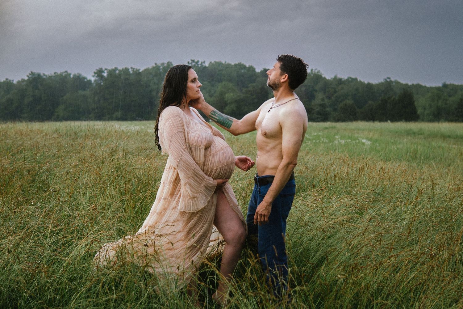 Couple stands lovingly together in a tall grass meadow during golden hour with stormy skies in the background.