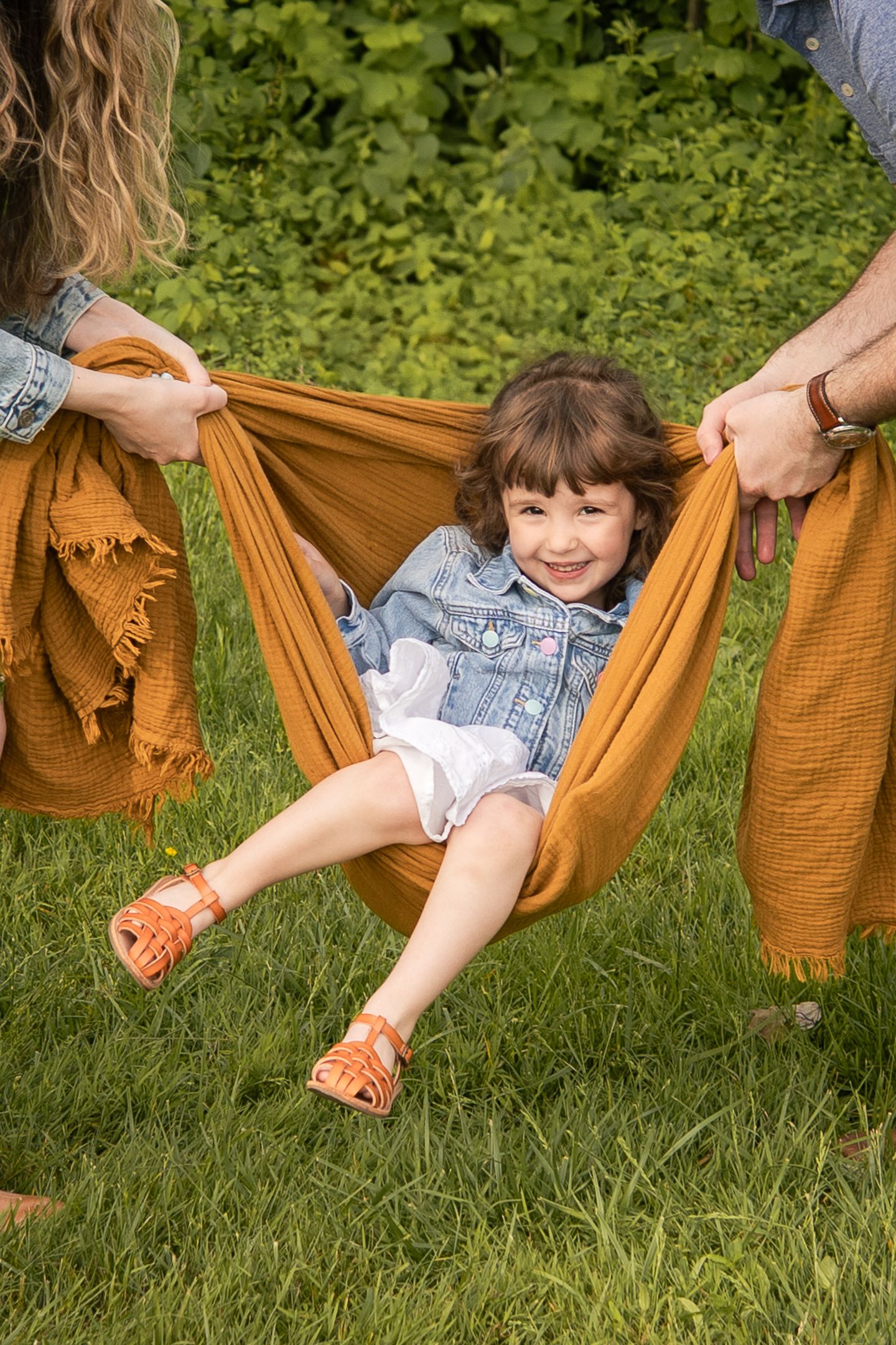 Two people swinging a child in a hammock-style swing made from brown fabric outside on green grass.