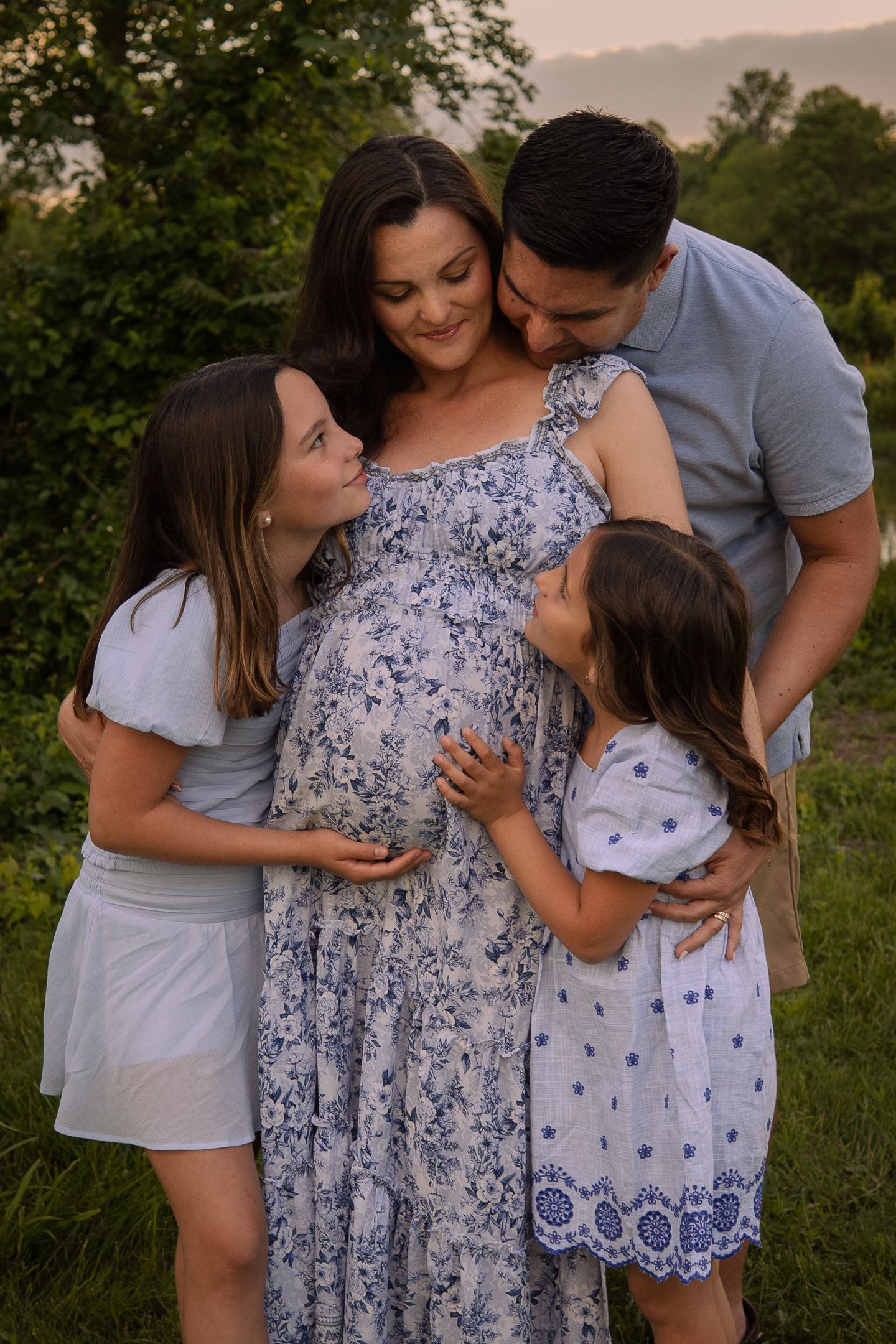 A loving family embraces a pregnant woman wearing a blue floral dress while standing in a grassy outdoor setting at sunset.