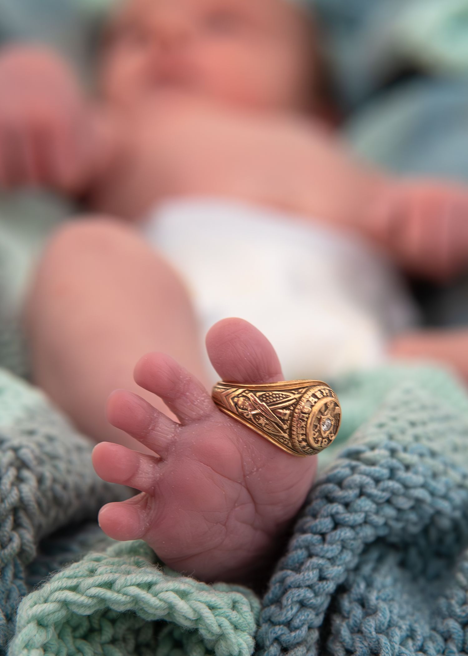 A tiny baby hand wearing a gold ring rests on a soft gray knitted blanket.