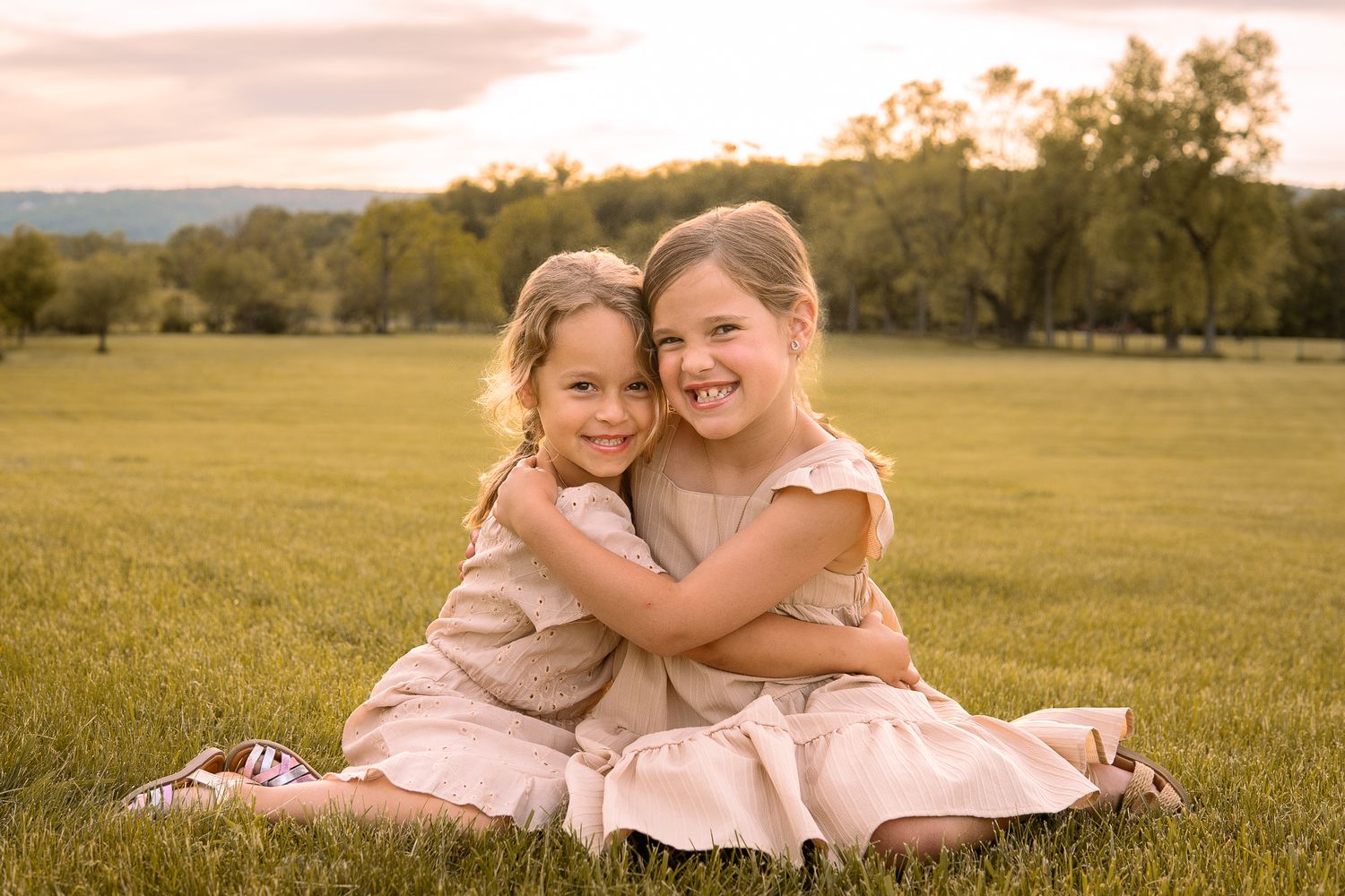 Two kids in pink dresses embrace while sitting in a grassy field at golden hour.