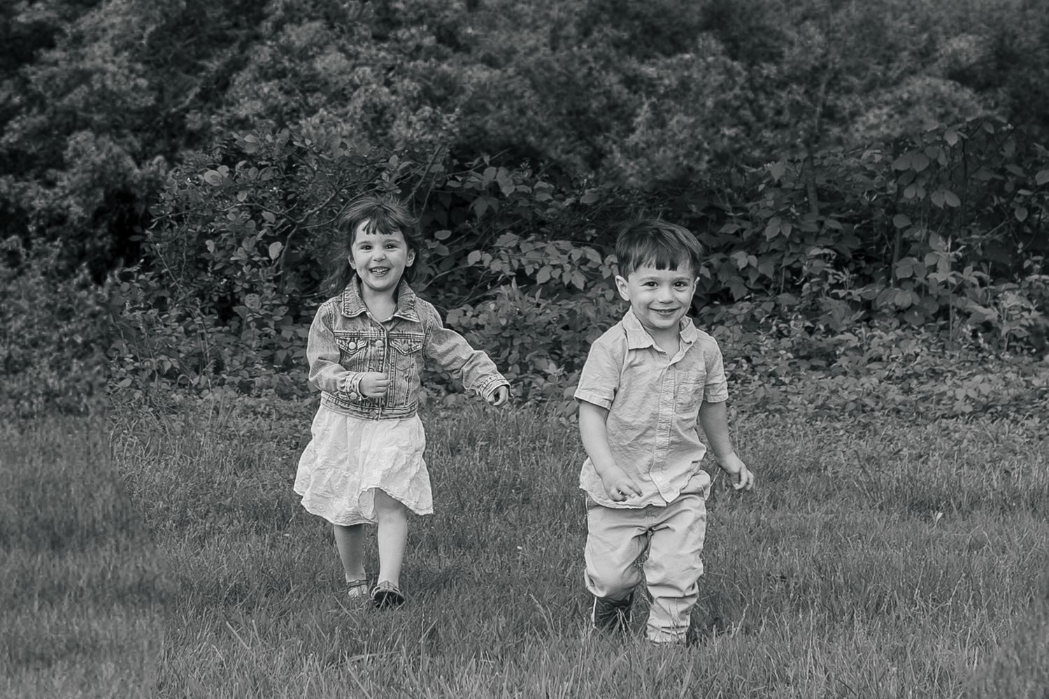 Black and white photo of two children running together in a grassy field with trees in the background.