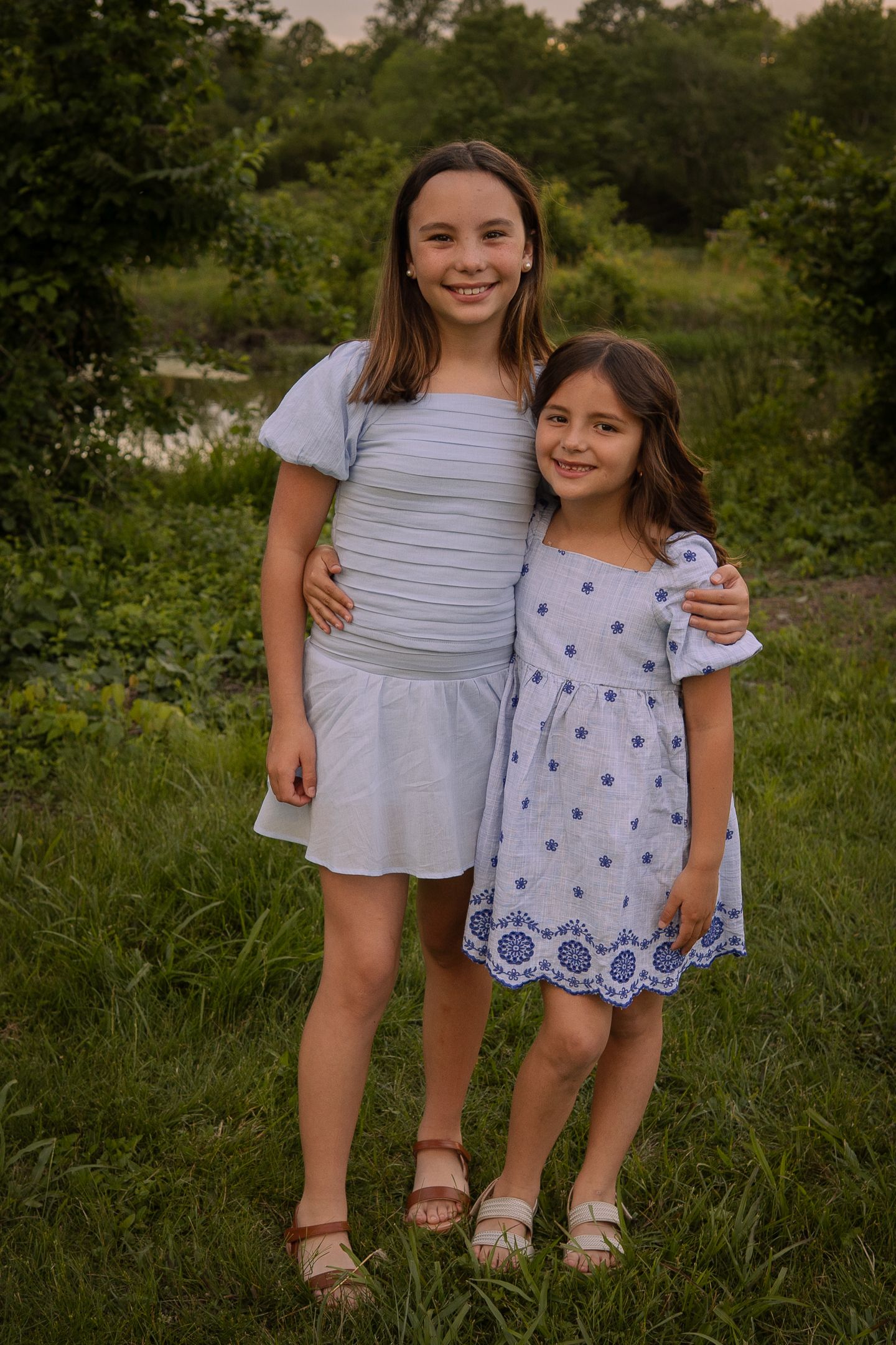 Two children wearing summer dresses stand together in a grassy outdoor setting at sunset.